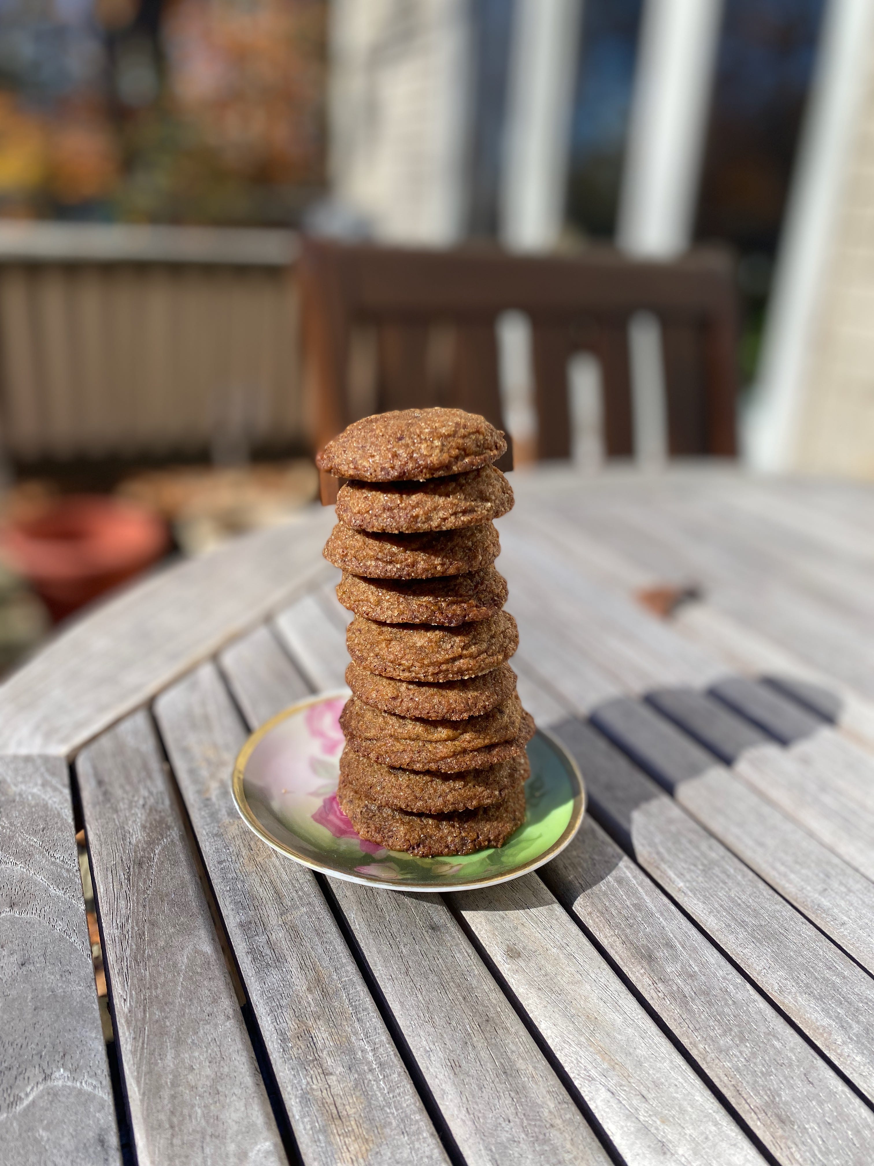 A tall stack of ten gingersnap cookies on a small hand-painted ceramic saucer sitting on a wooden porch table.