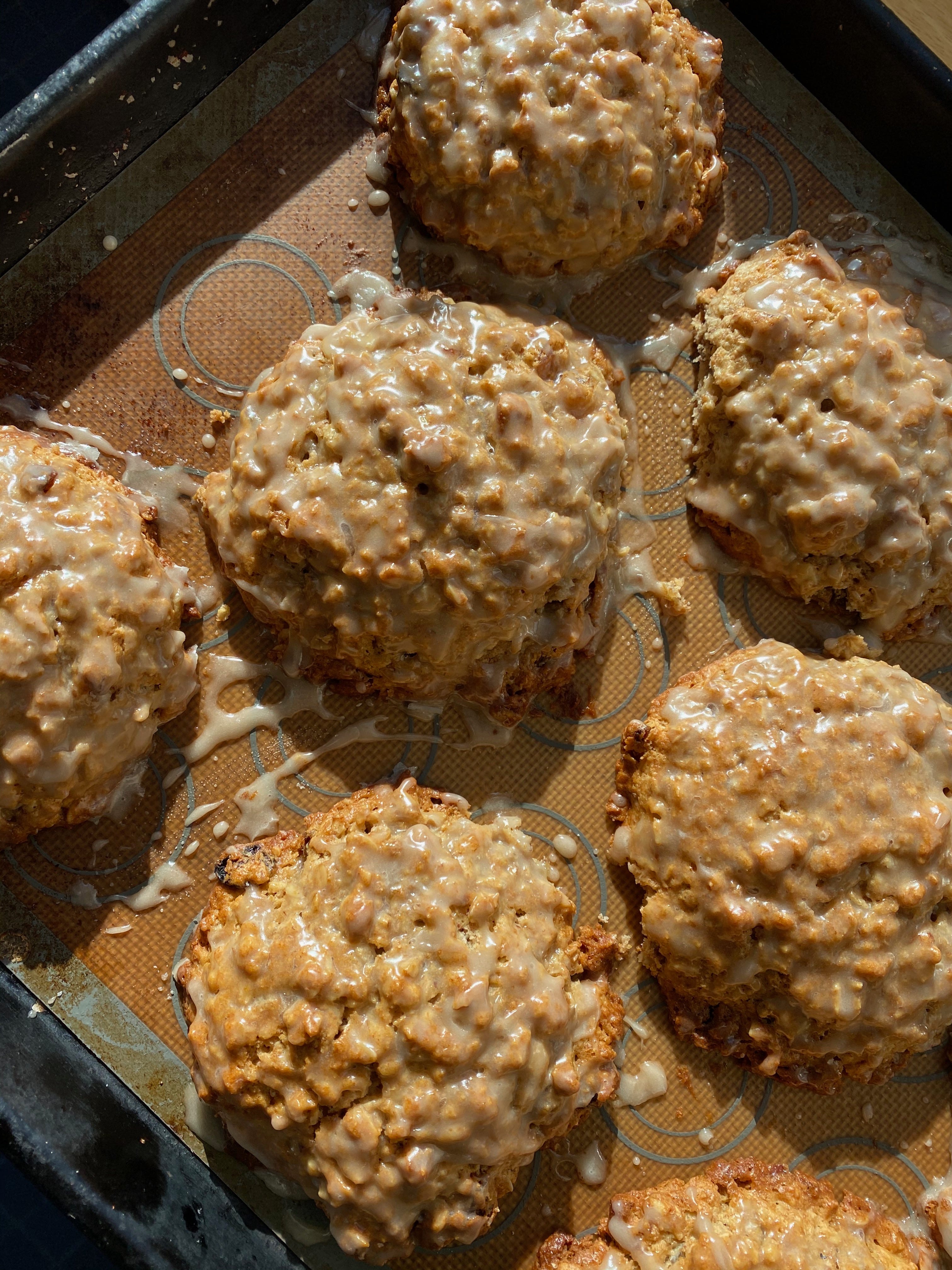 Six scones on a baking tray on a sunlit counter. They are round and craggy, topped with a thin white glaze.