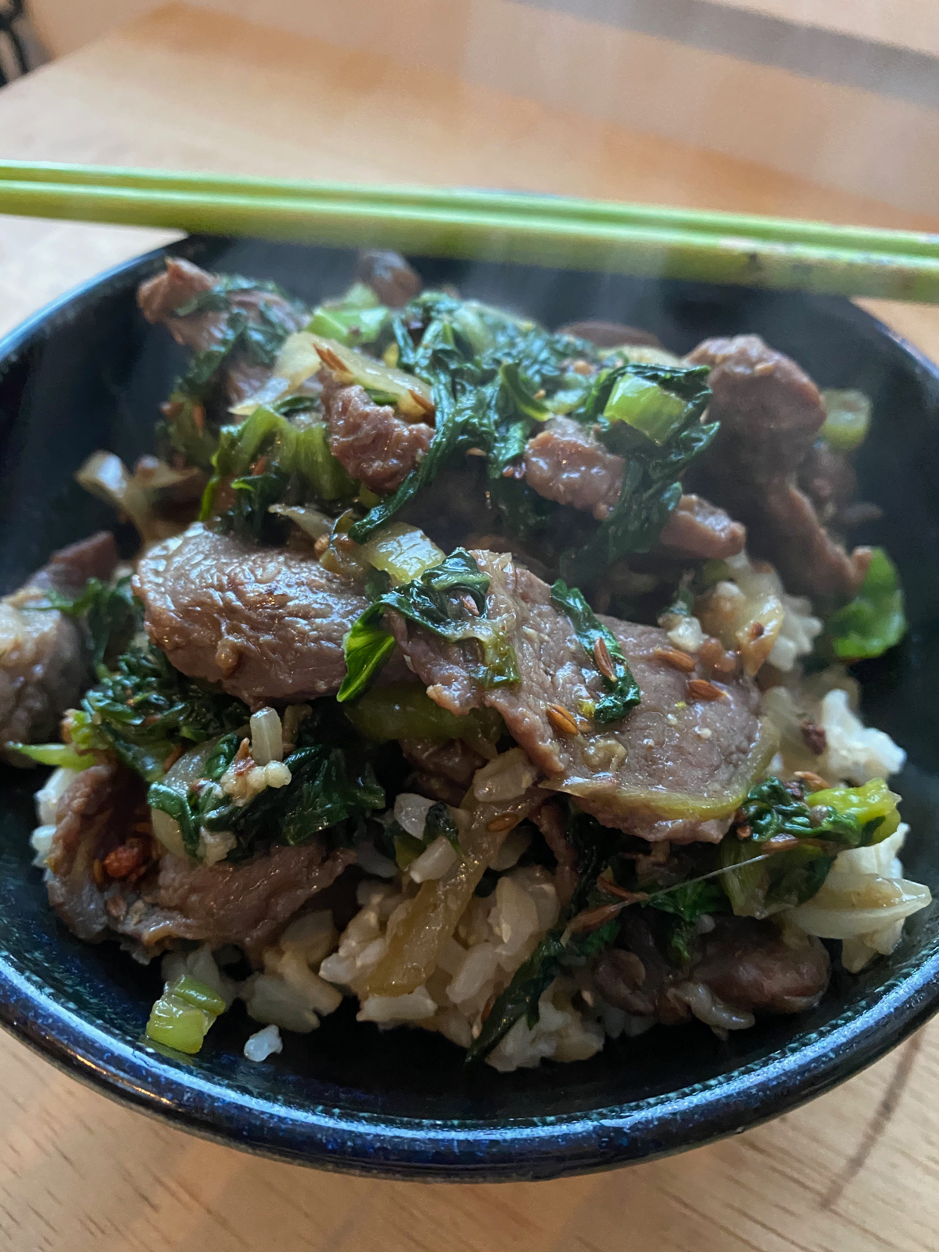 A blue ceramic bowl of lamb sir-fry, containing pieces of lamb, slivers of boy choy, rice, and cumin seeds. A pair of green chopsticks rests on the edge of the bowl.
