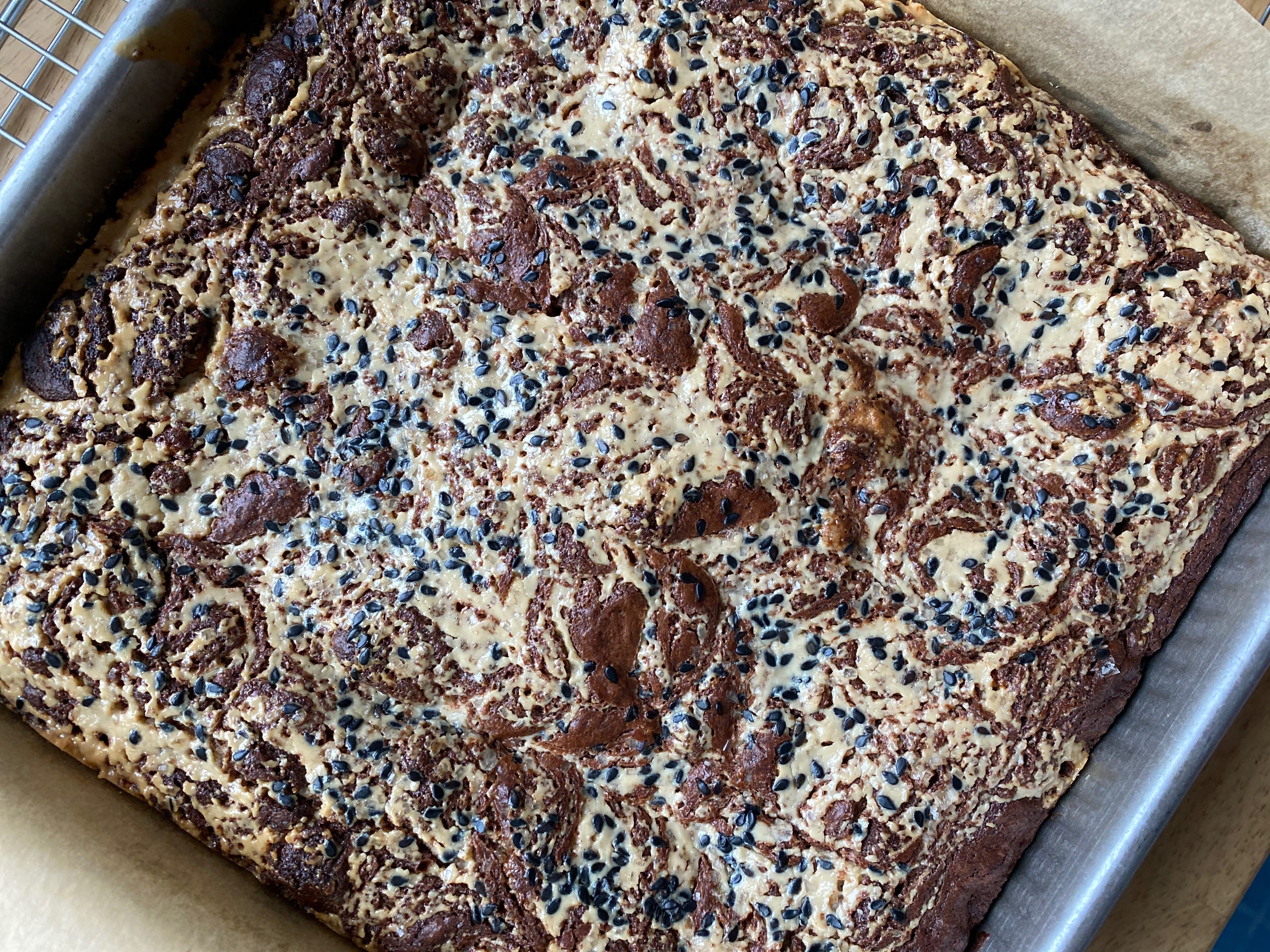 A pan of tahini brownies on a wooden counter. The tahini makes a swirly marbled pattern, and black sesame seeds are scattered across the top.