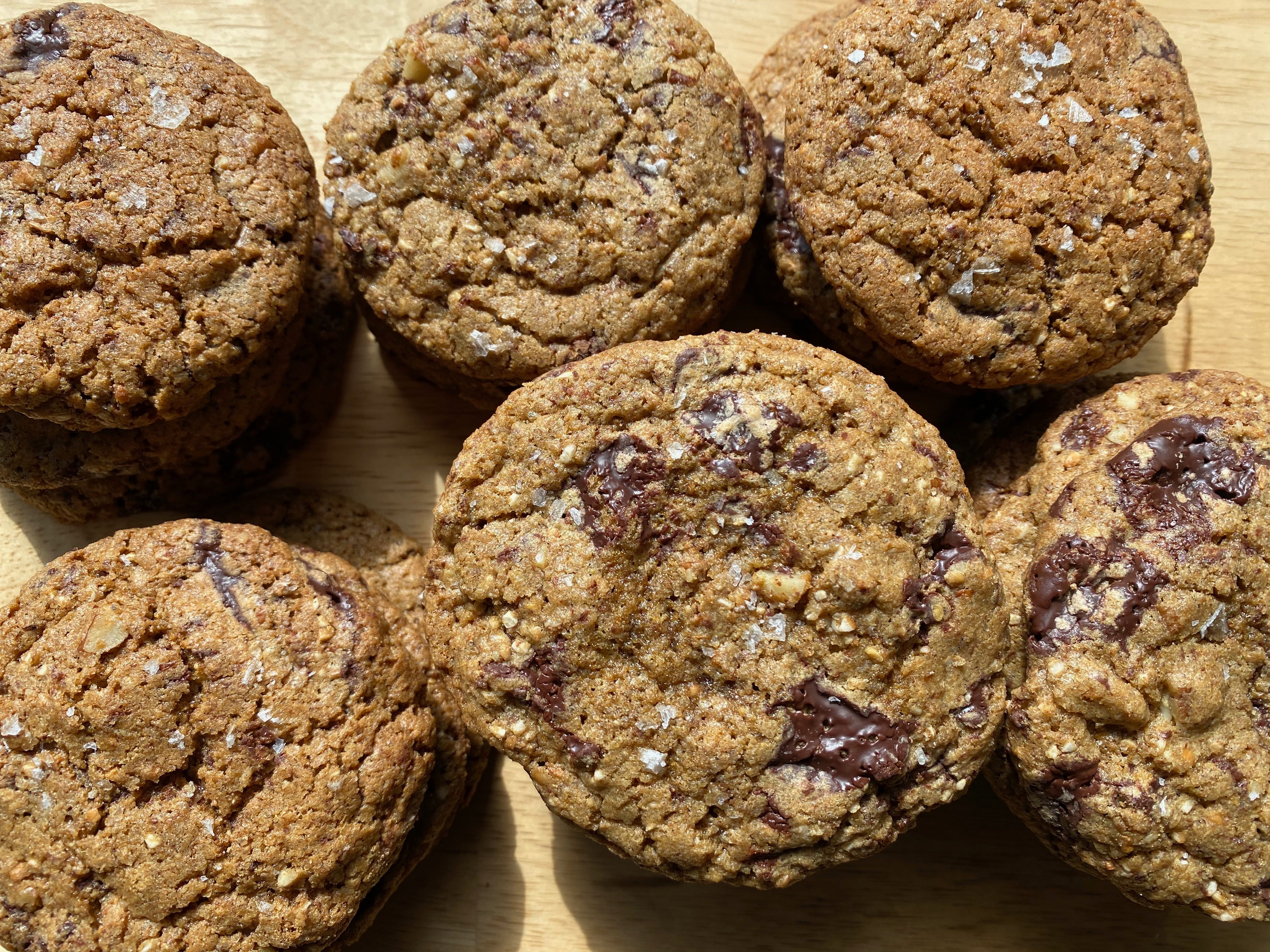 Stacks of multigrain chocolate chip cookies on a wooden surface. There are six stacks arranged in two rows, and you can only see the top cookies. The cookies are golden brown circles with crinkled tops studded with big chunks of chocolate and flaky salt.