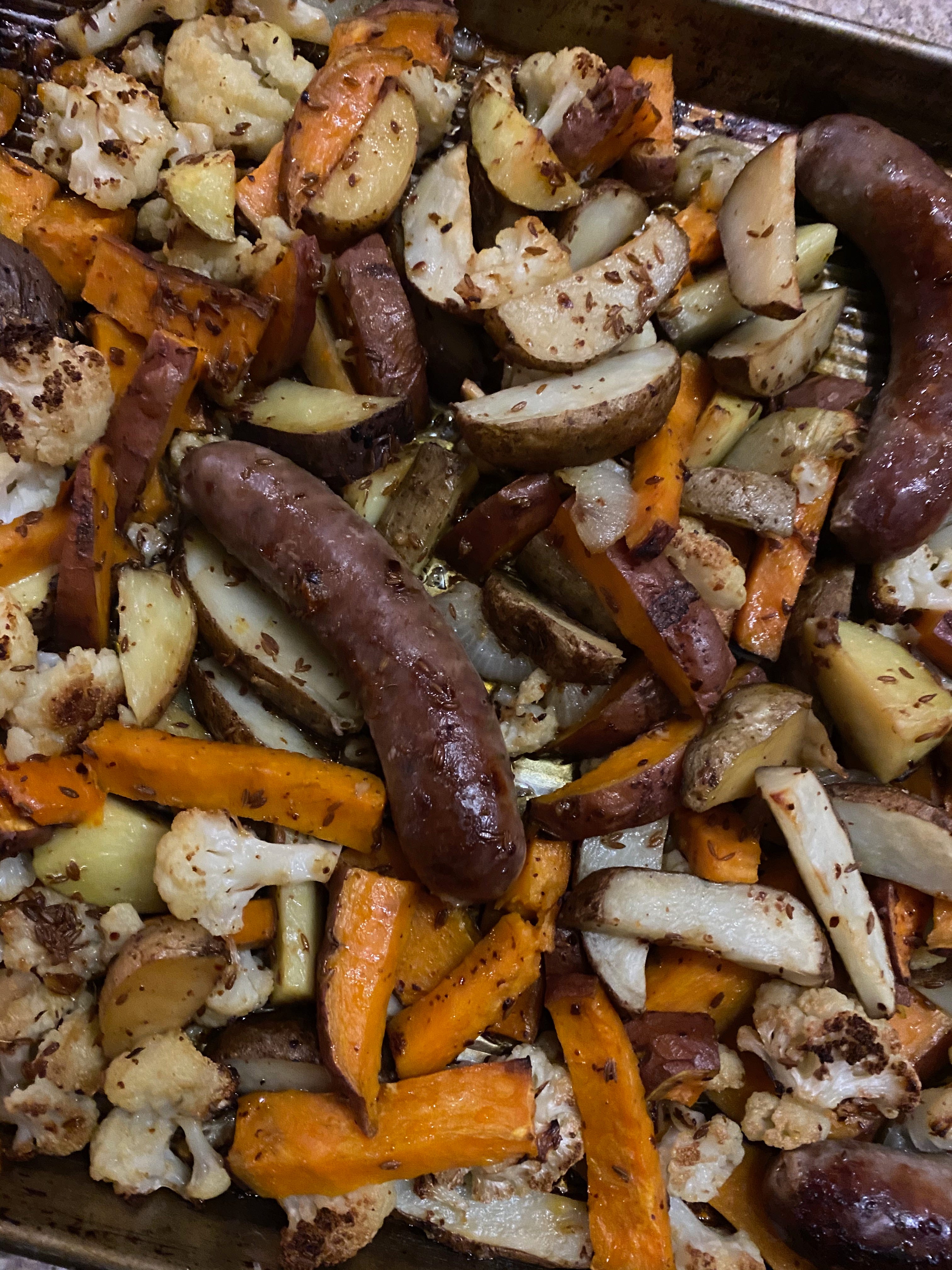 A closeup of a tray of roasted vegetables: sweet potatoes, white potatoes, cauliflower florets, and sausage links. The veggies are browned around the edges and covered in flecks of cumin seeds.
