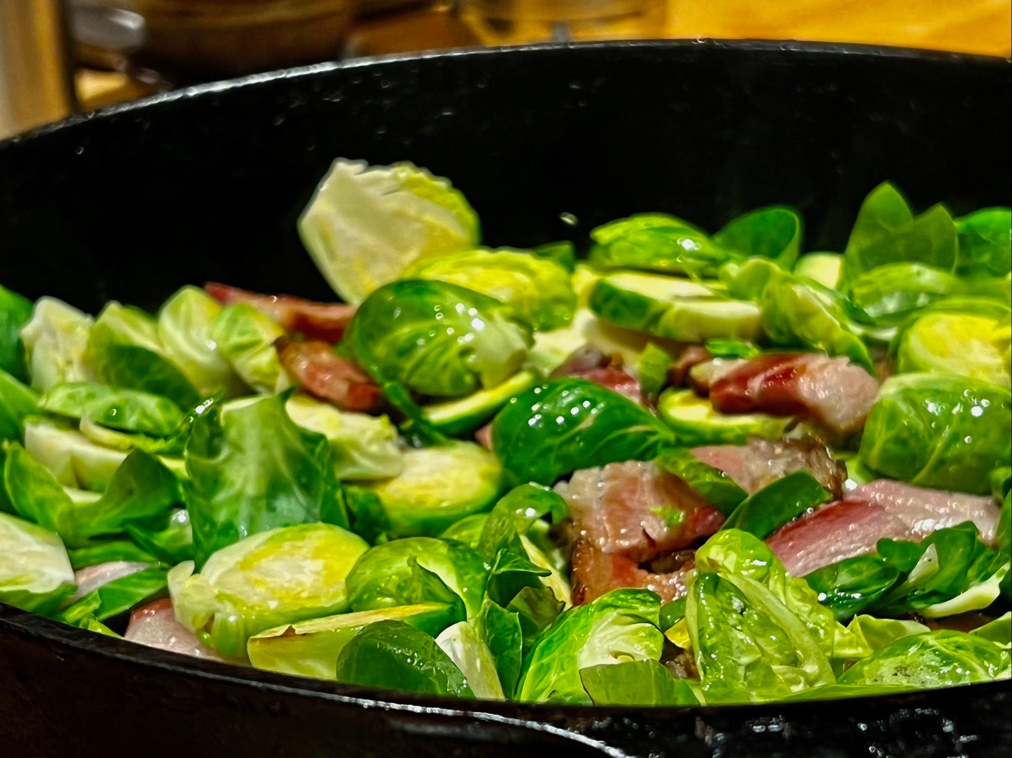 A cast iron pan of bright green brussel sprouts, sliced into rounds, cooking with small strips of bacon.
