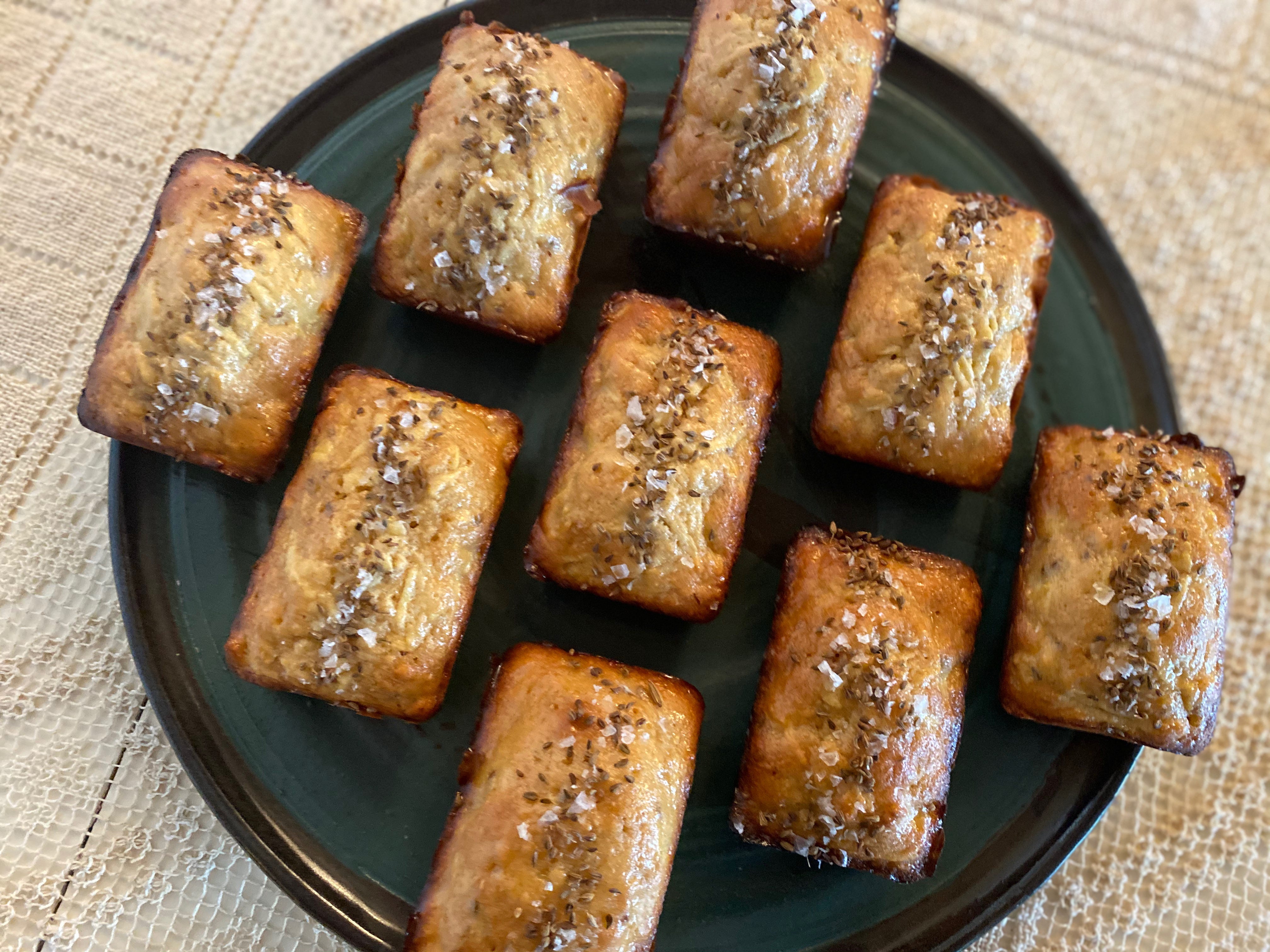 Nine small rectangular loaf cakes sit on a round green ceramic plate. The cakes are arranged in a diamond pattern. They are golden brown and each one has a vertical line of seeds and flaky salt running down its center. The plate is on a lace tablecloth.