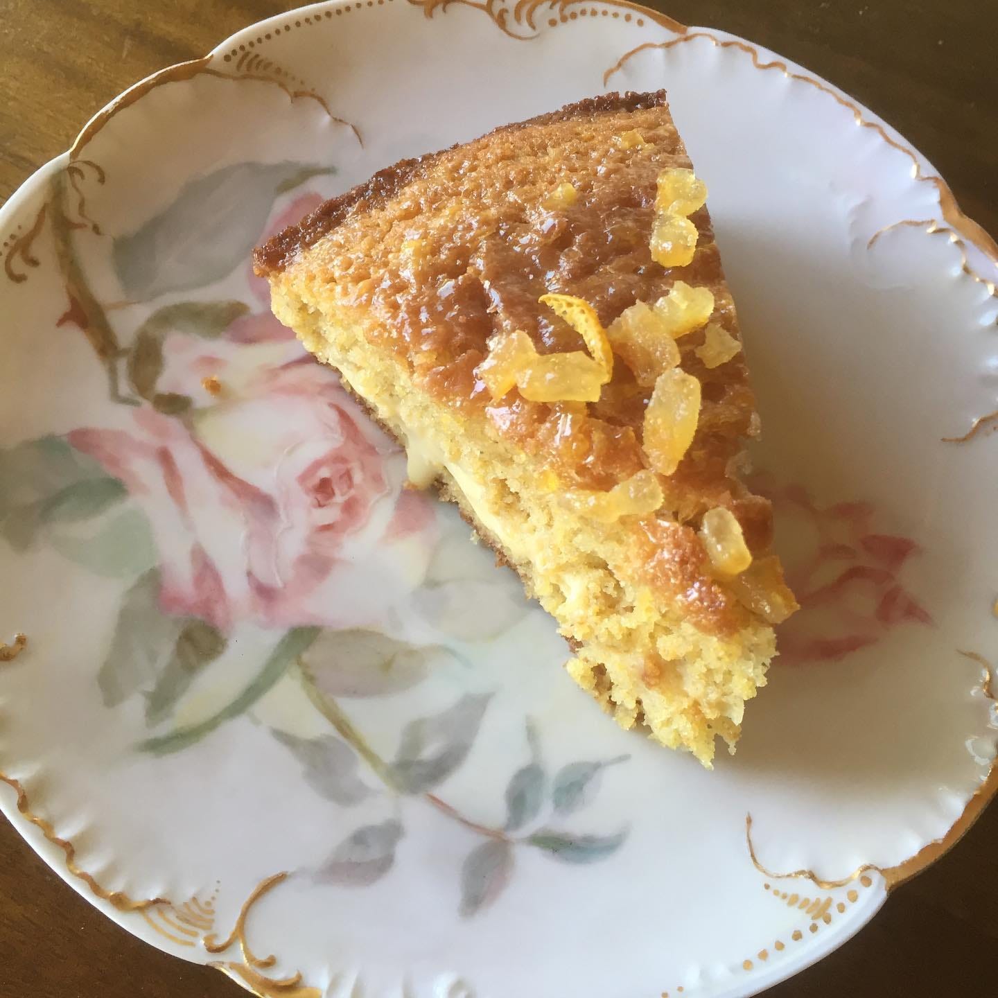 A slice of golden-brown cake on a small ceramic plate painted with flowers. Orange curd oozes out from between the layers, and the top is sprinkled with candied orange peel.