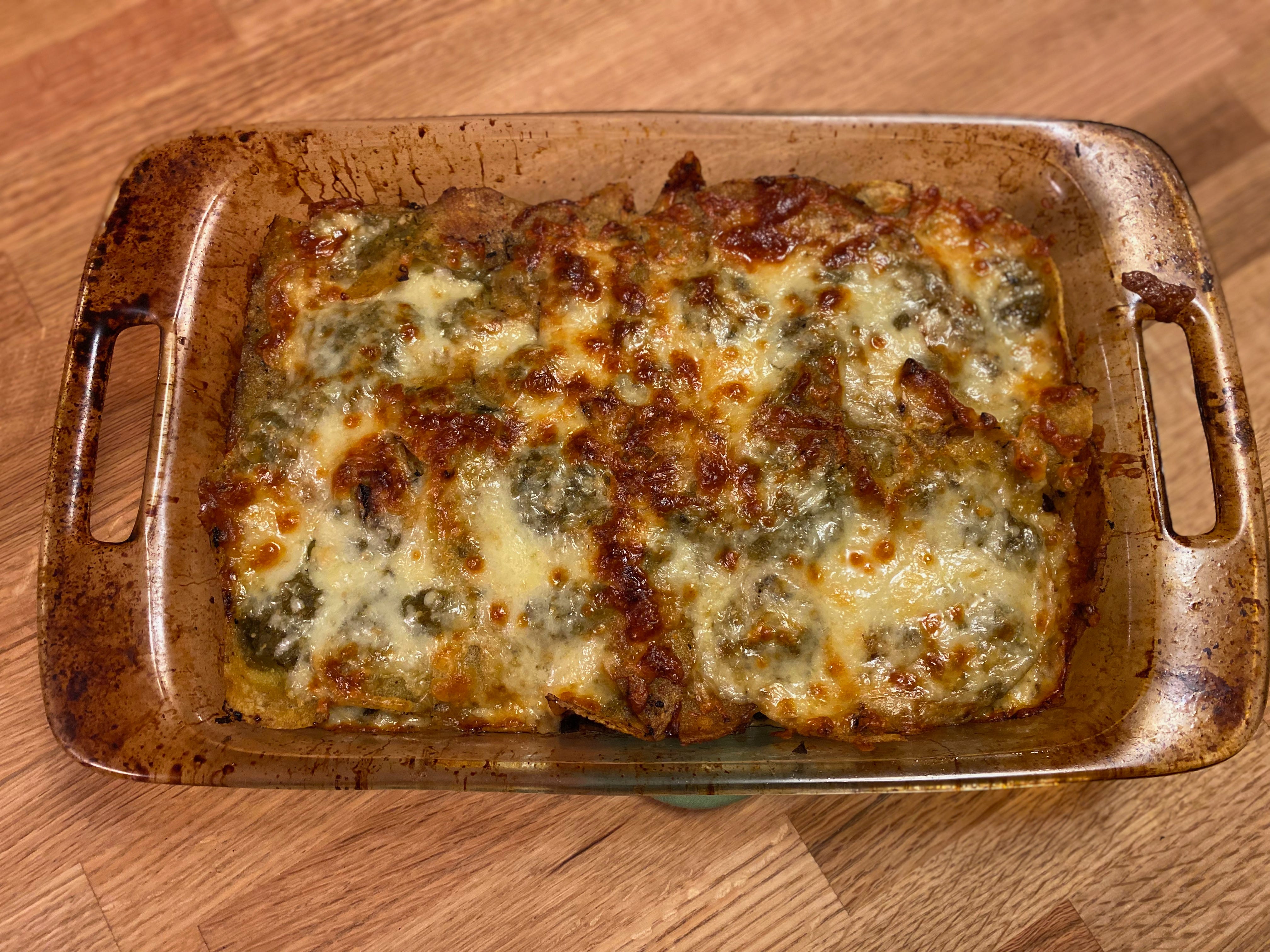 A glass casserole dish on a wooden counter. The casserole is topped with gooey, melty, bubbling cheese, browned in places. Green tomatillo salsa peeks out from the layers beneath.