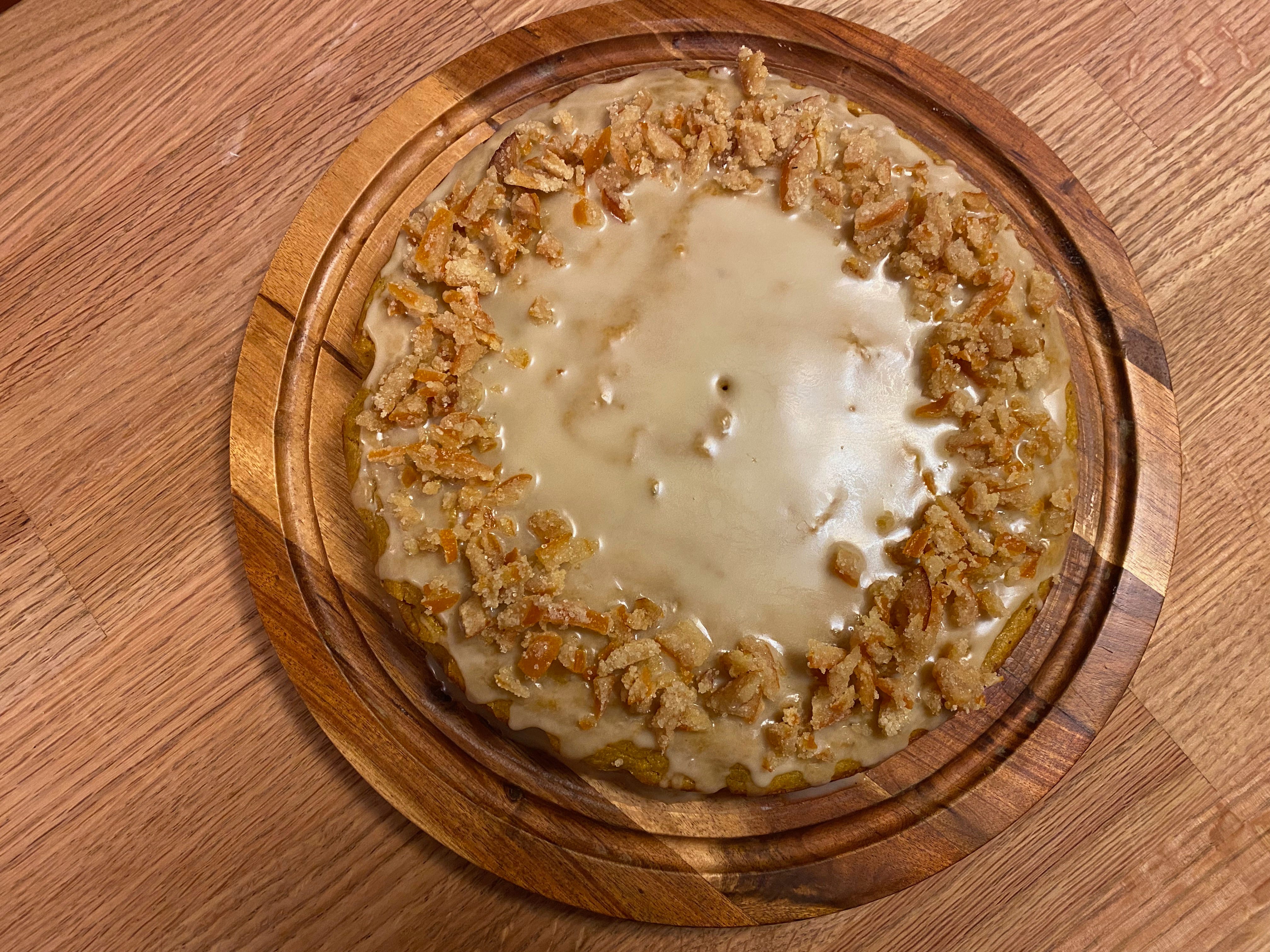 A round cake topped with a shiny glaze and chopped candied orange peel sits on a wooden cake plate on a counter.