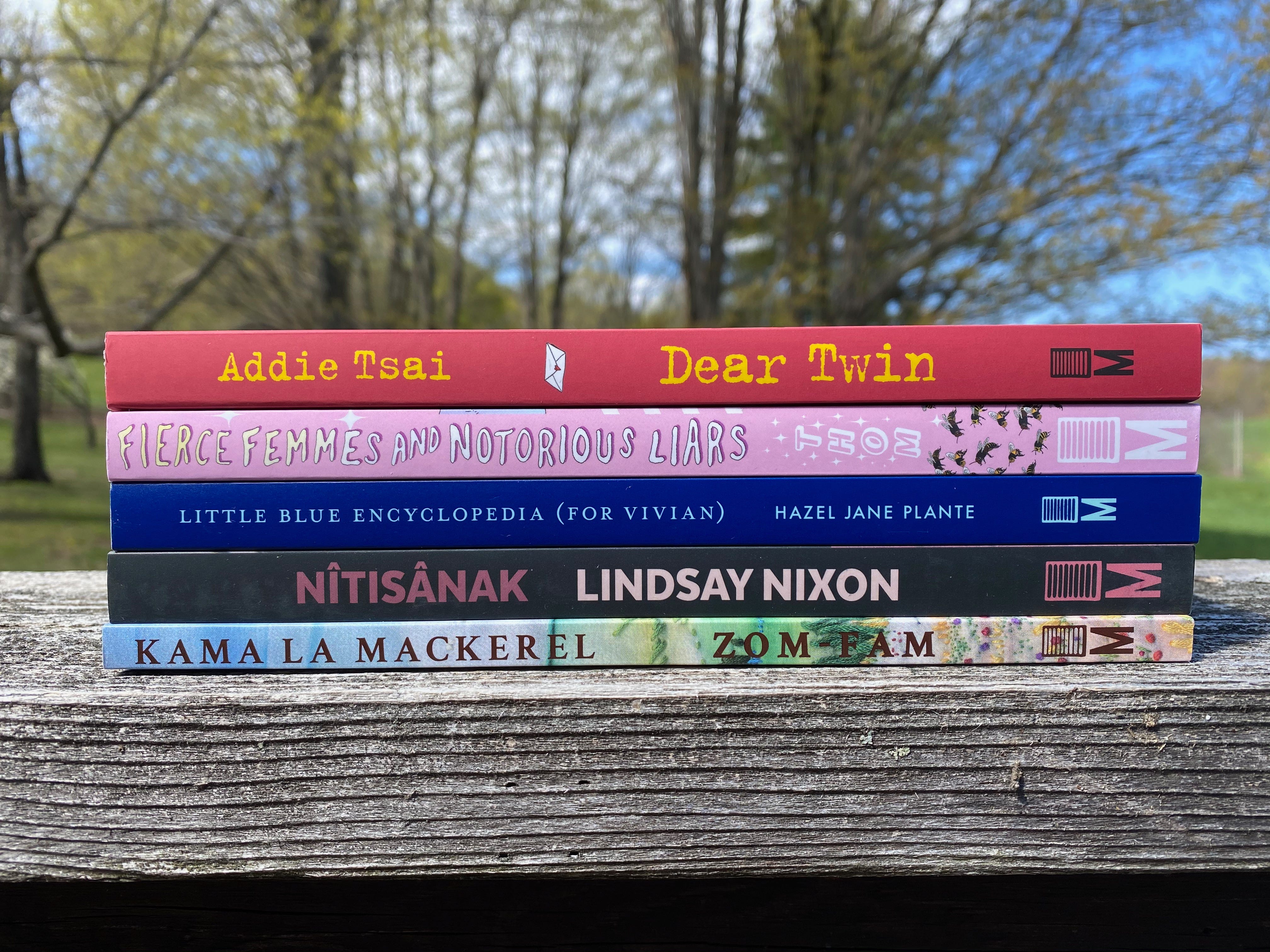 A stack of books on a wooden railing in front of a view of trees and sky. The books: Zom-Fam by Kamala Mackerel, Nîtisânak by Lindsay Nixon, Little Blue Encyclopedia (for Vivian) by Hazel Jane Plante, Fierce Femmes and Notorious Liars by Kai Cheng Thom, and Dear Twin by Addie Tsai.