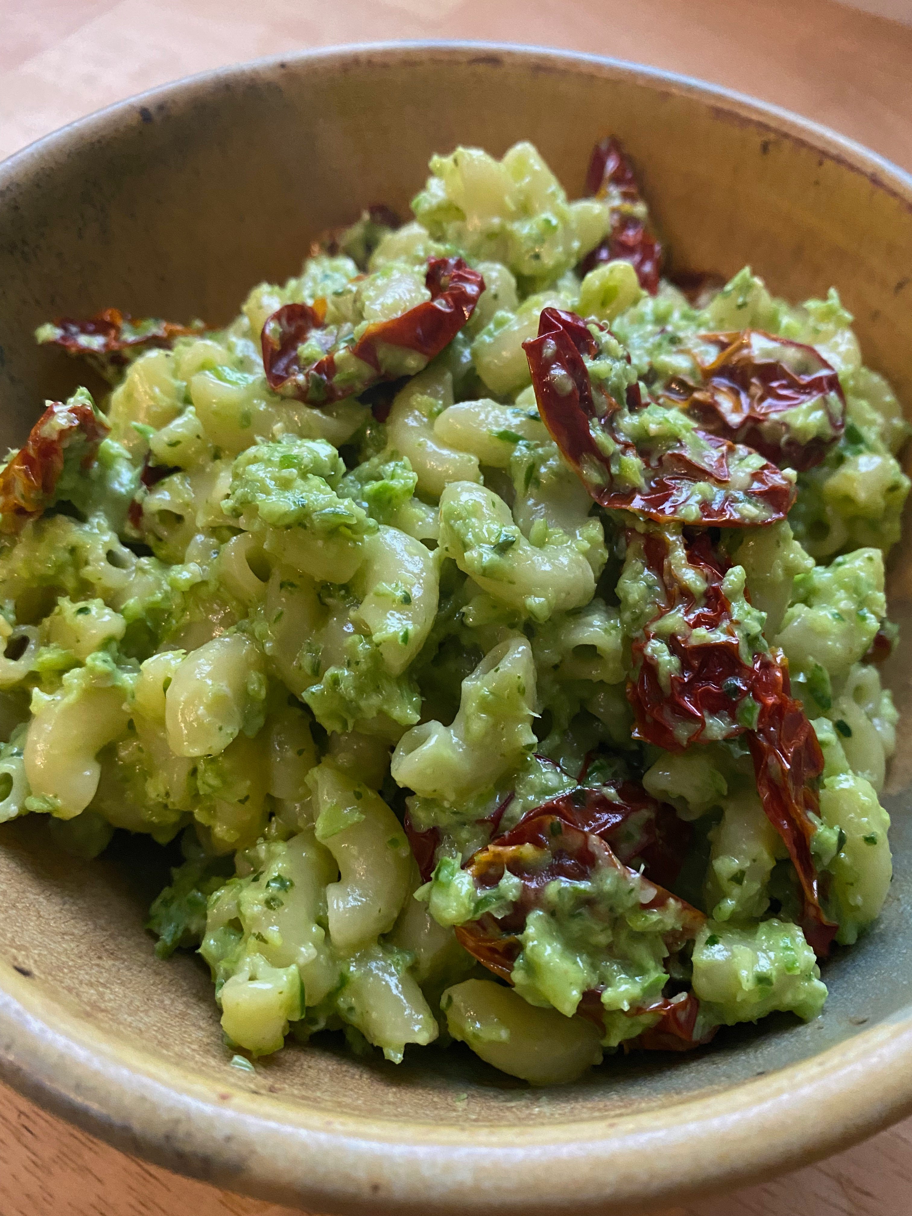 Closeup of a bowl of small elbow noodles coated in garlic scape pesto, topped with wrinkly red sun-dried tomatoes.