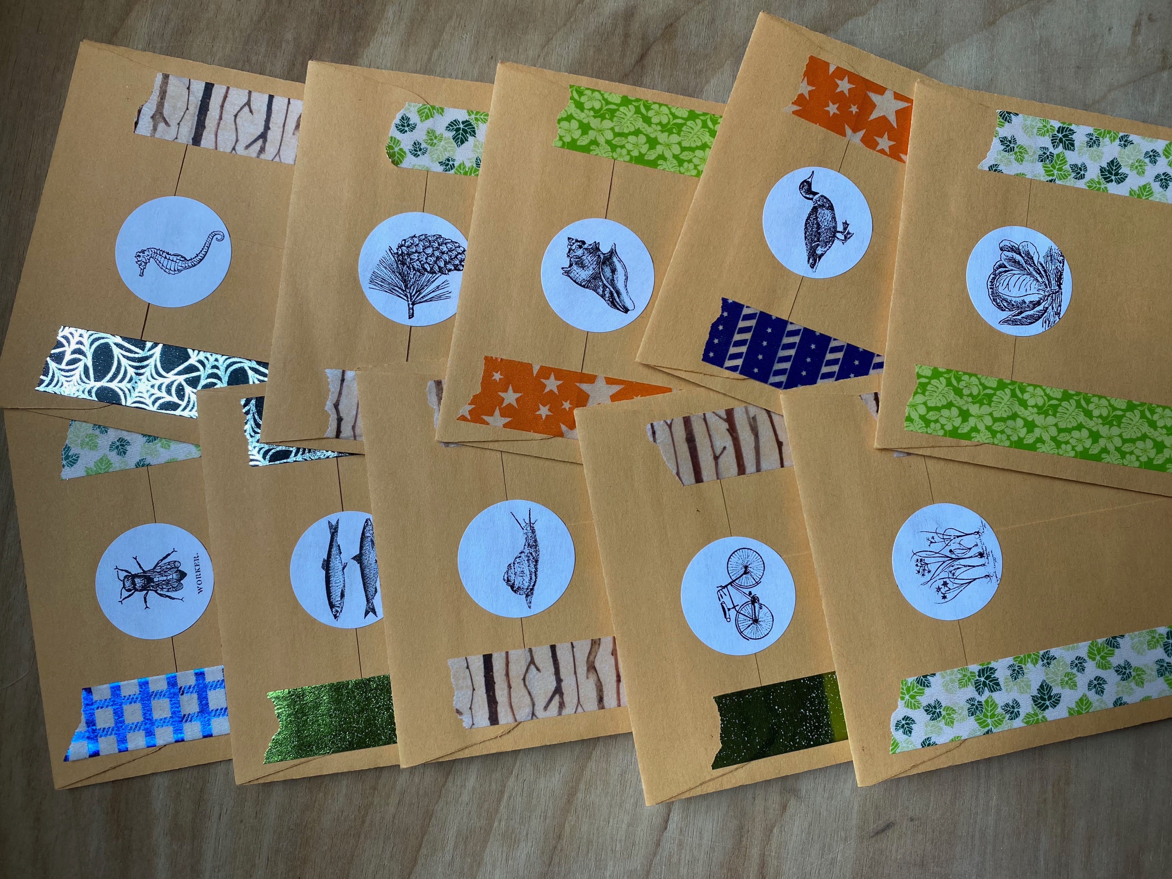Ten small brown envelopes in two rows of five, overlapping. Each one is sealed by a round sticker with a different design (seahorse, pine cone, cabbage, bike, snail, fish, flowers, duck, bee, shell). They are decorated with colorful washi tape in various styles and patterns—leaves, branches, stars, spiderwebs, stripes.