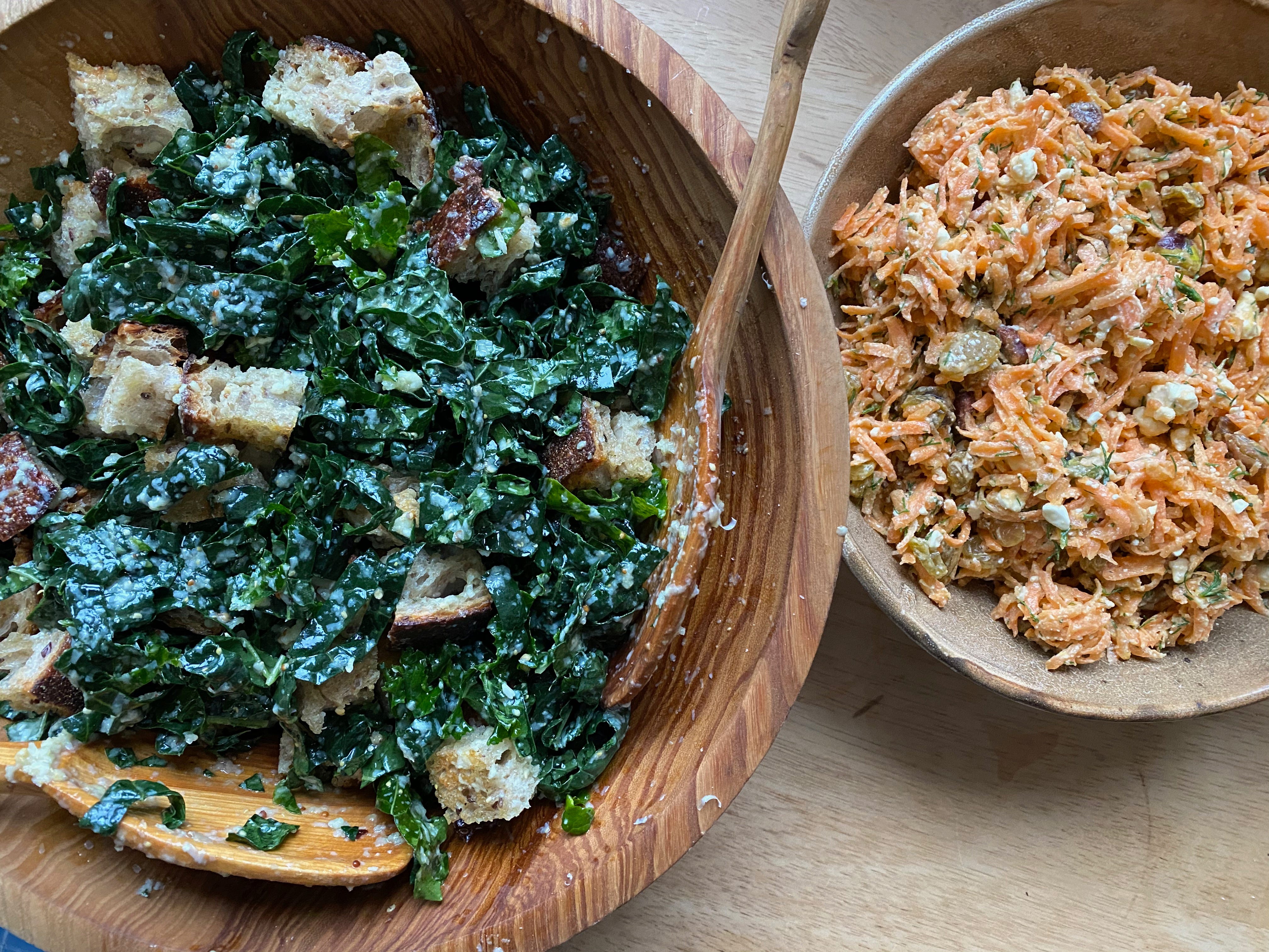 Close up of two bowls on a wooden surface. A large wooden bowl holds kale salad with croutons. A smaller ceramic bowl contains a grated carrot salad studded with raisins, dill, and chunks of feta.