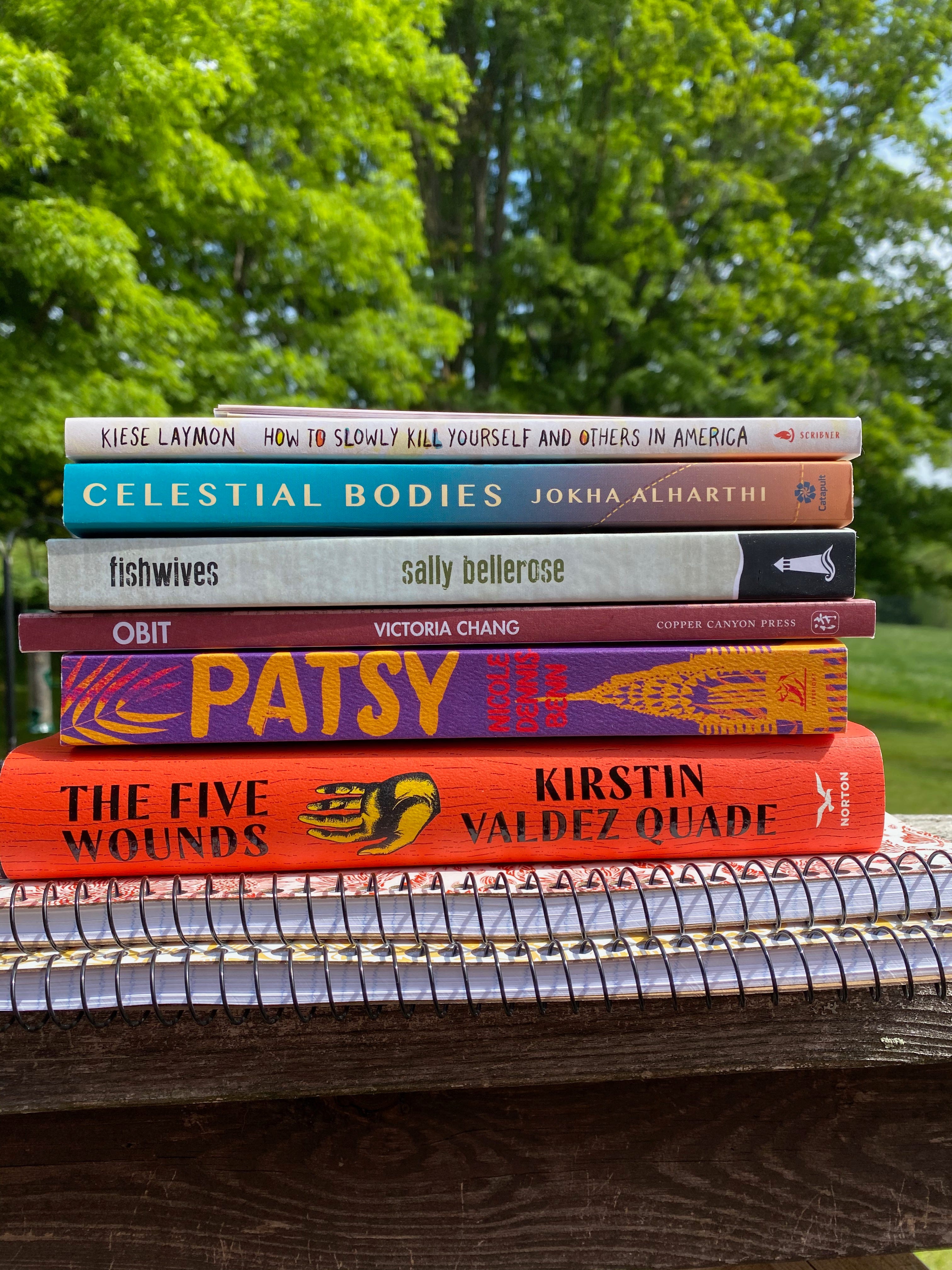 A stack of books (and two spiral notebooks) on a wooden railing in front of green trees. The books are: The Five Wounds, Patsy, Celestial Bodies, Obit, Fishwives, and How to Slowly Kill Yourself and Others in America.
