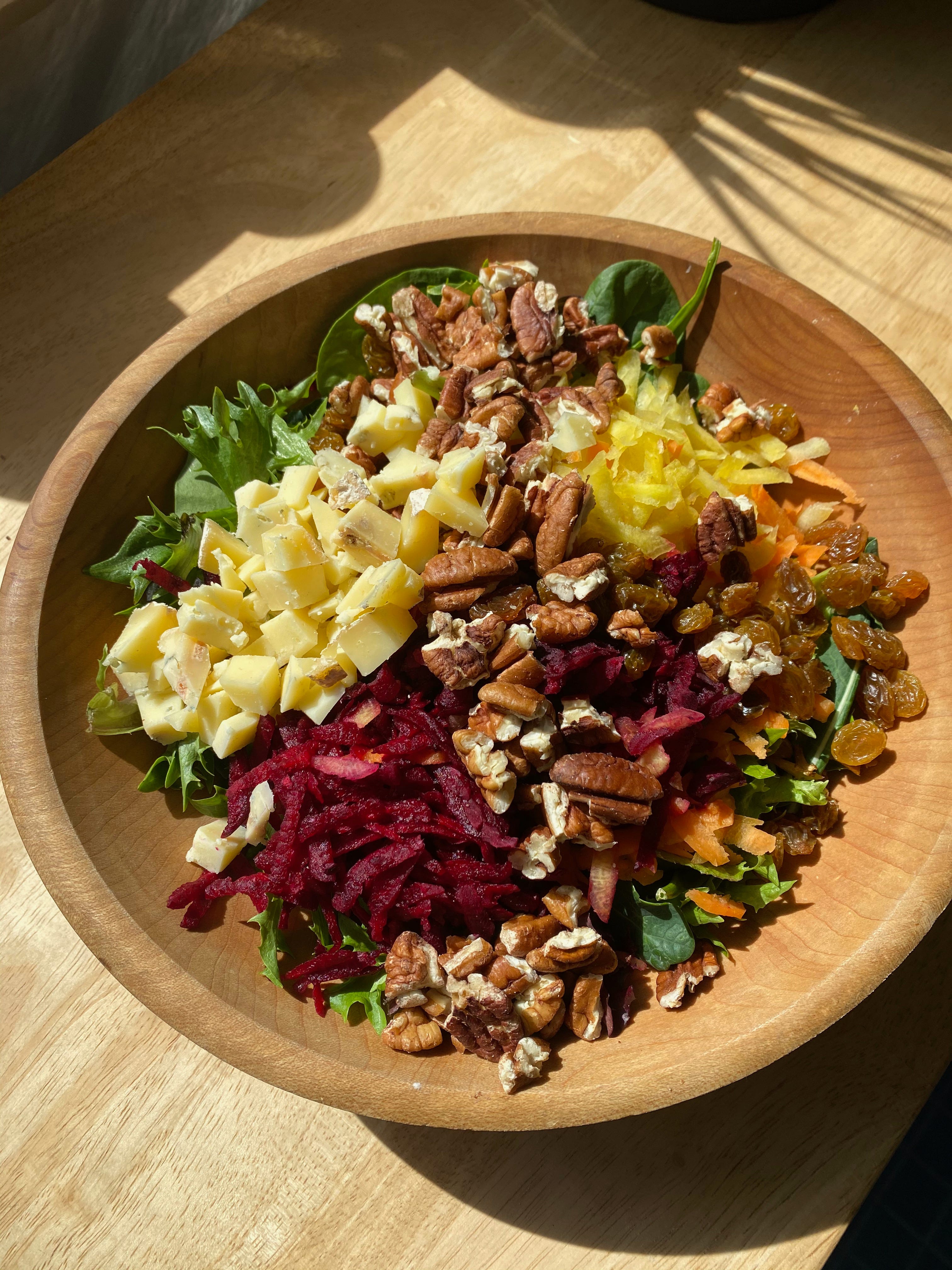 A wooden bowl of salad sits on a sunlit wooden counter. The salad is not tossed. On top of the greens, you can see piles of cubed cheese, grated red beet, grated yellow carrot, golden raisins, and chopped pecans.