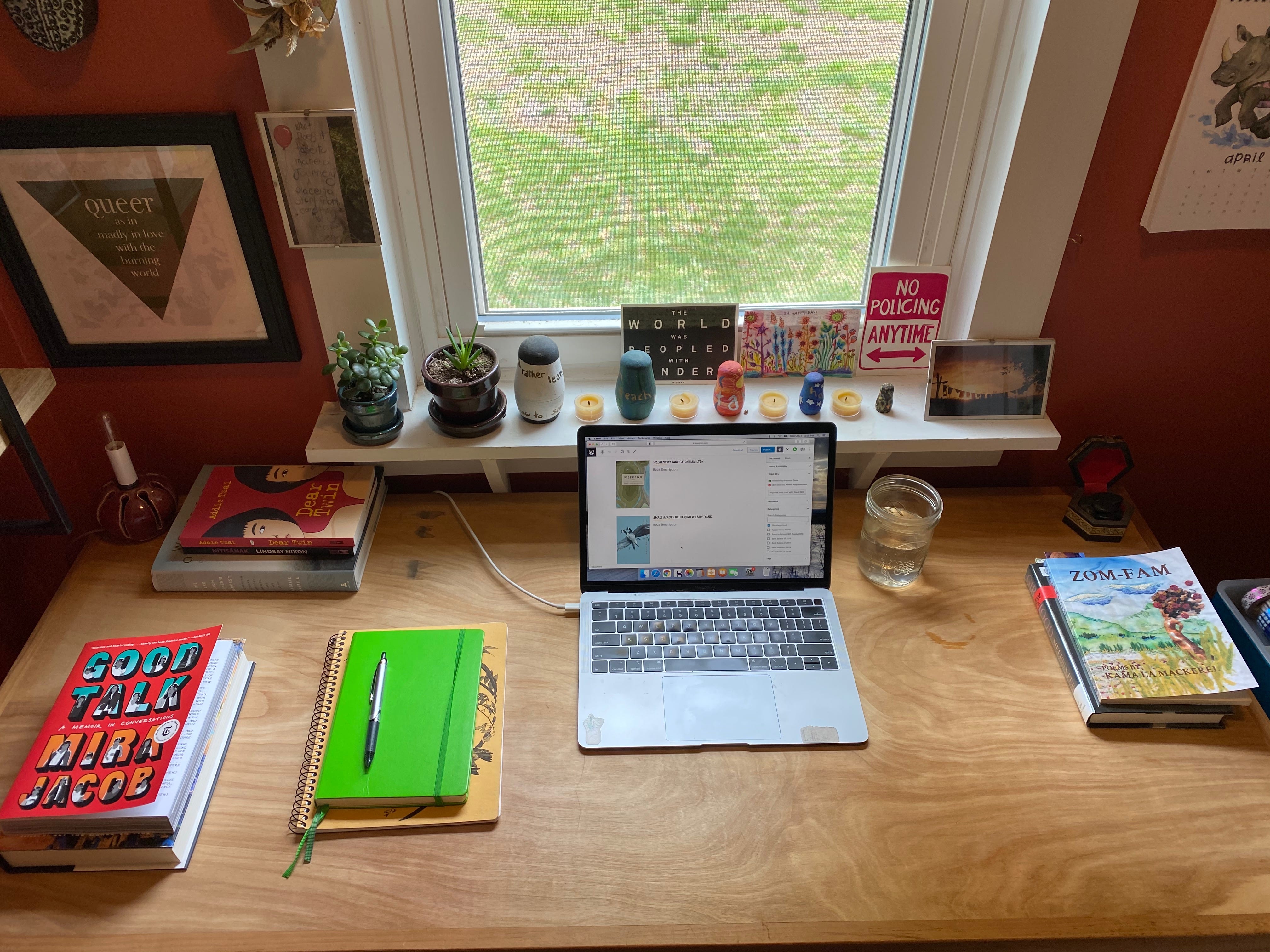 View of a wooden desk from above. There’s an open laptop, several stacks of books, and a stack of notebooks with a pen on top. You can see grass out the window, and the windowsill holds several small succulents, postcards, painted figurines, and candles.
