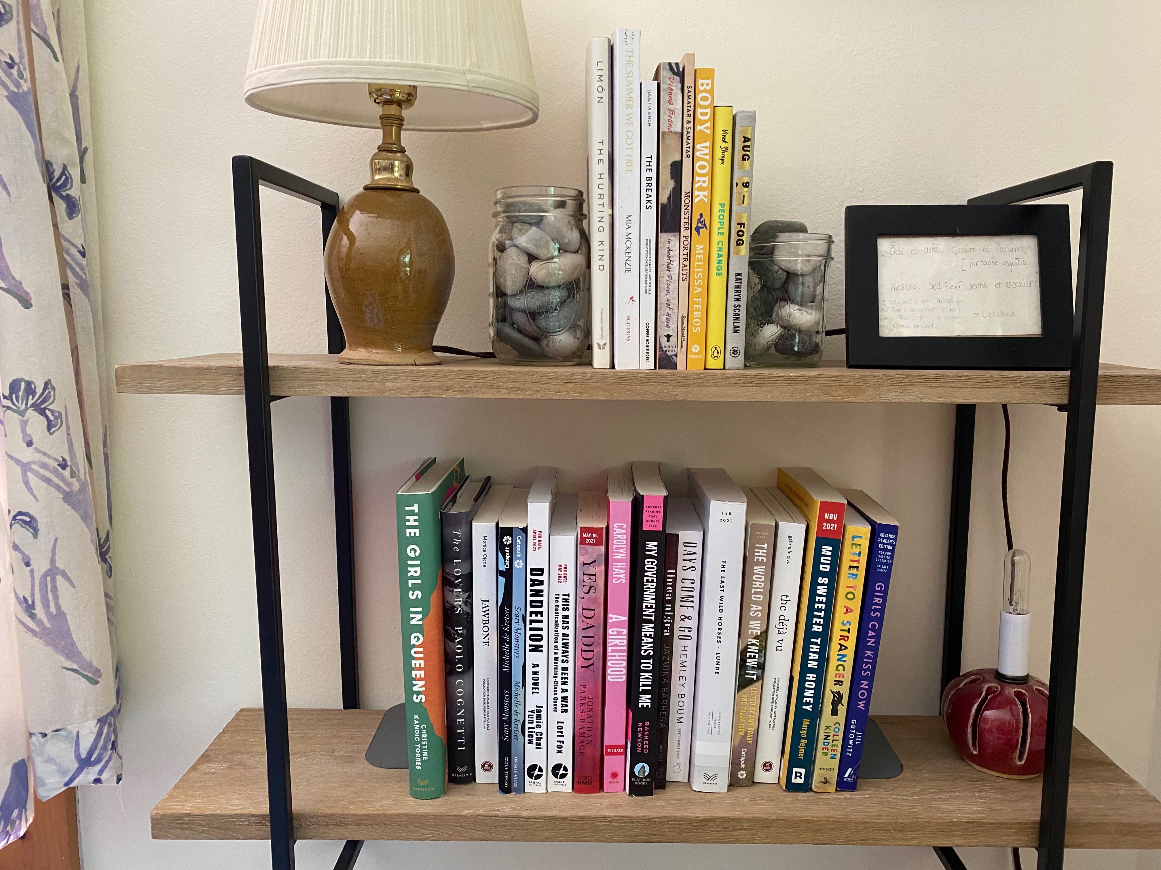 A wall mounted shelf displaying two shelves of books. The books on the top shelf are bookended by two mason jars of stones, and it also holds a small ceramic lamp and a small black frame of the Catullus quote.