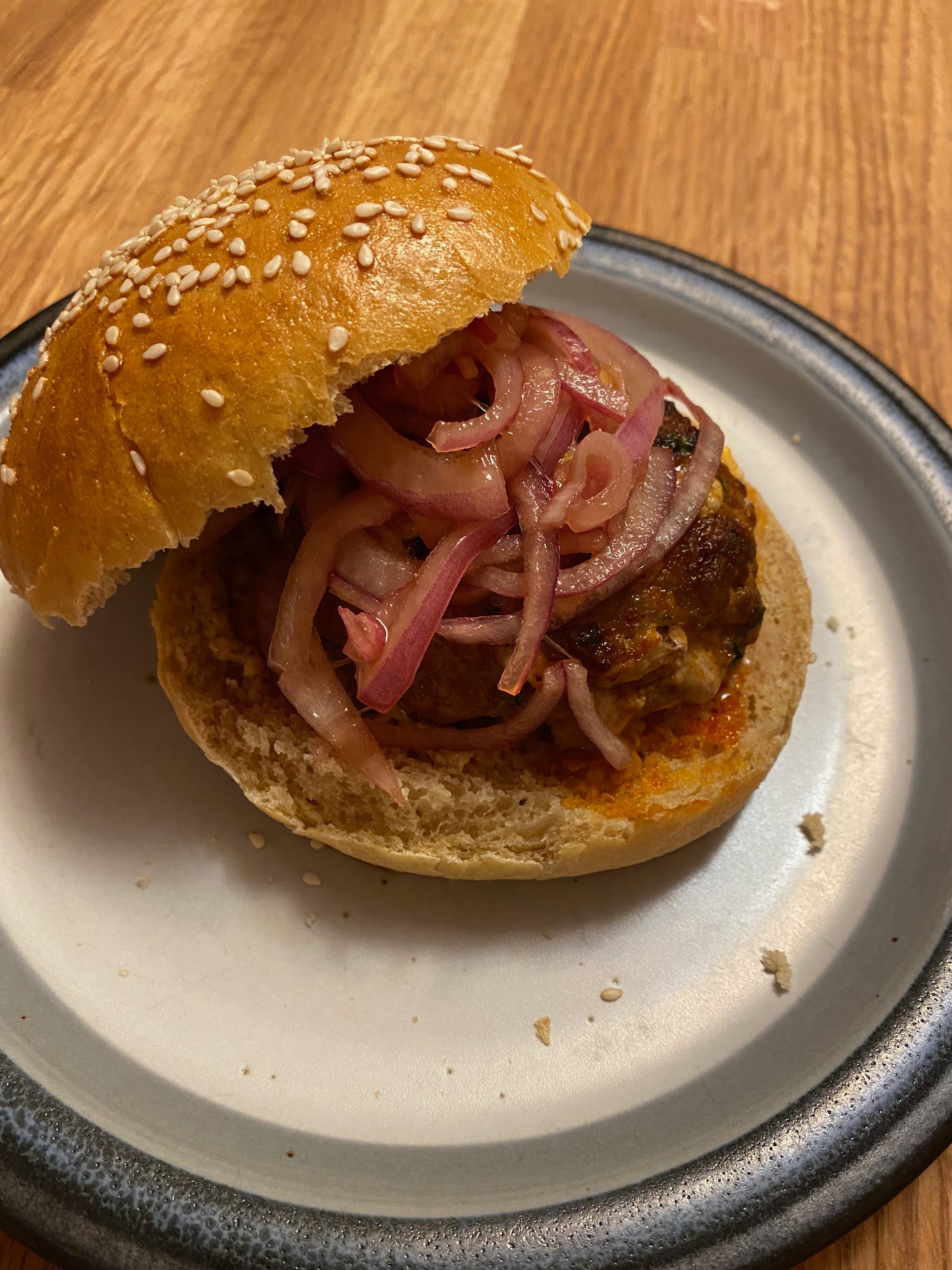 A burger on a blue ceramic plate. The top of the bun sits to one side, revealing a burger patty topped with a pile of pickled onions inside.