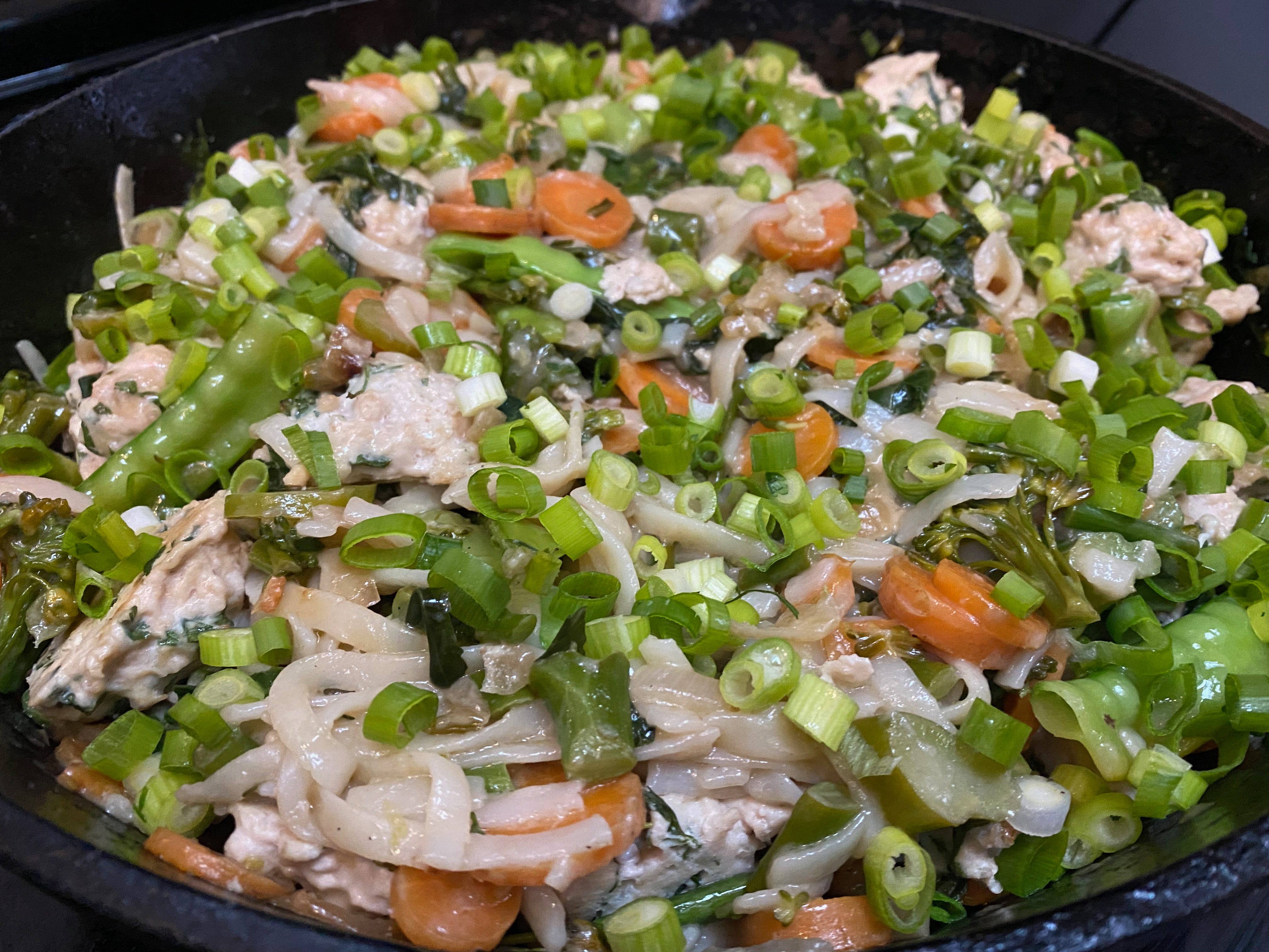 Closeup of a frying pan full of rice noodles and veggies, topped with lots of minced scallions, orange carrot rounds, and snow peas.
