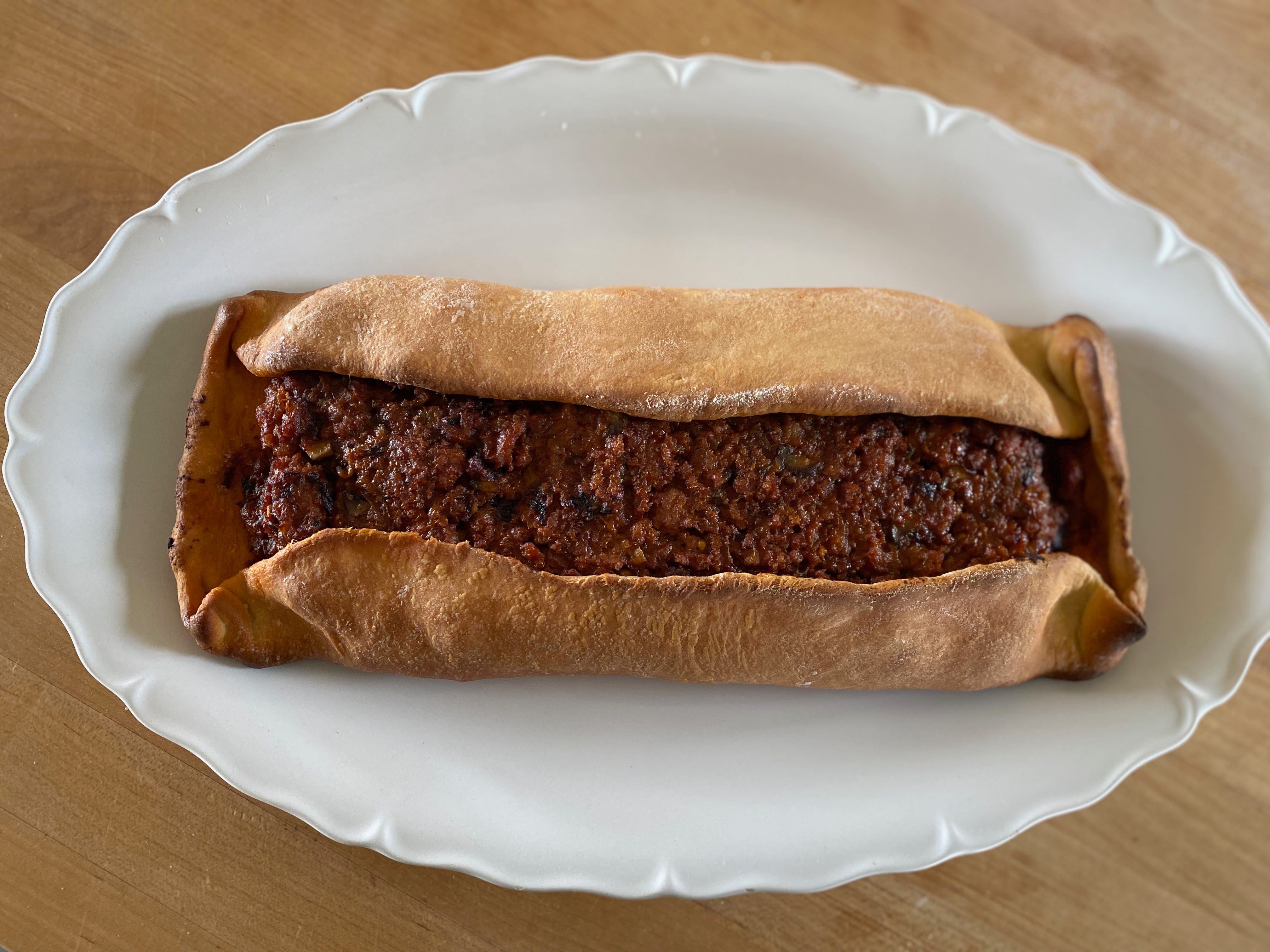 A golden brown, rectangular flatbread roll, with pork filling visible in the center, sits on a large white oval platter on a kitchen counter.