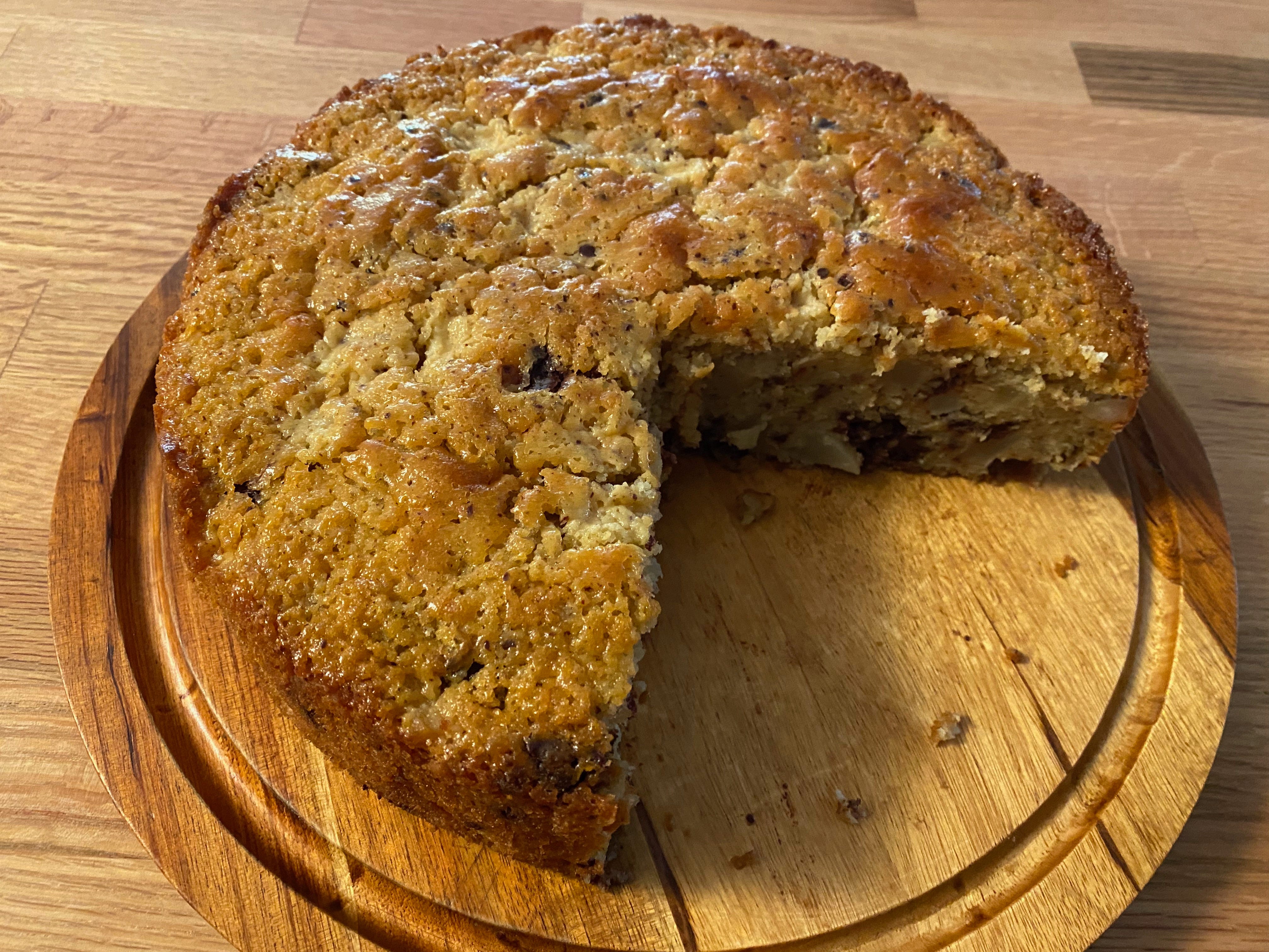 A round cake with a large piece cut out of it sits on a wooden board. The top of the cake is golden brown and wrinkly.