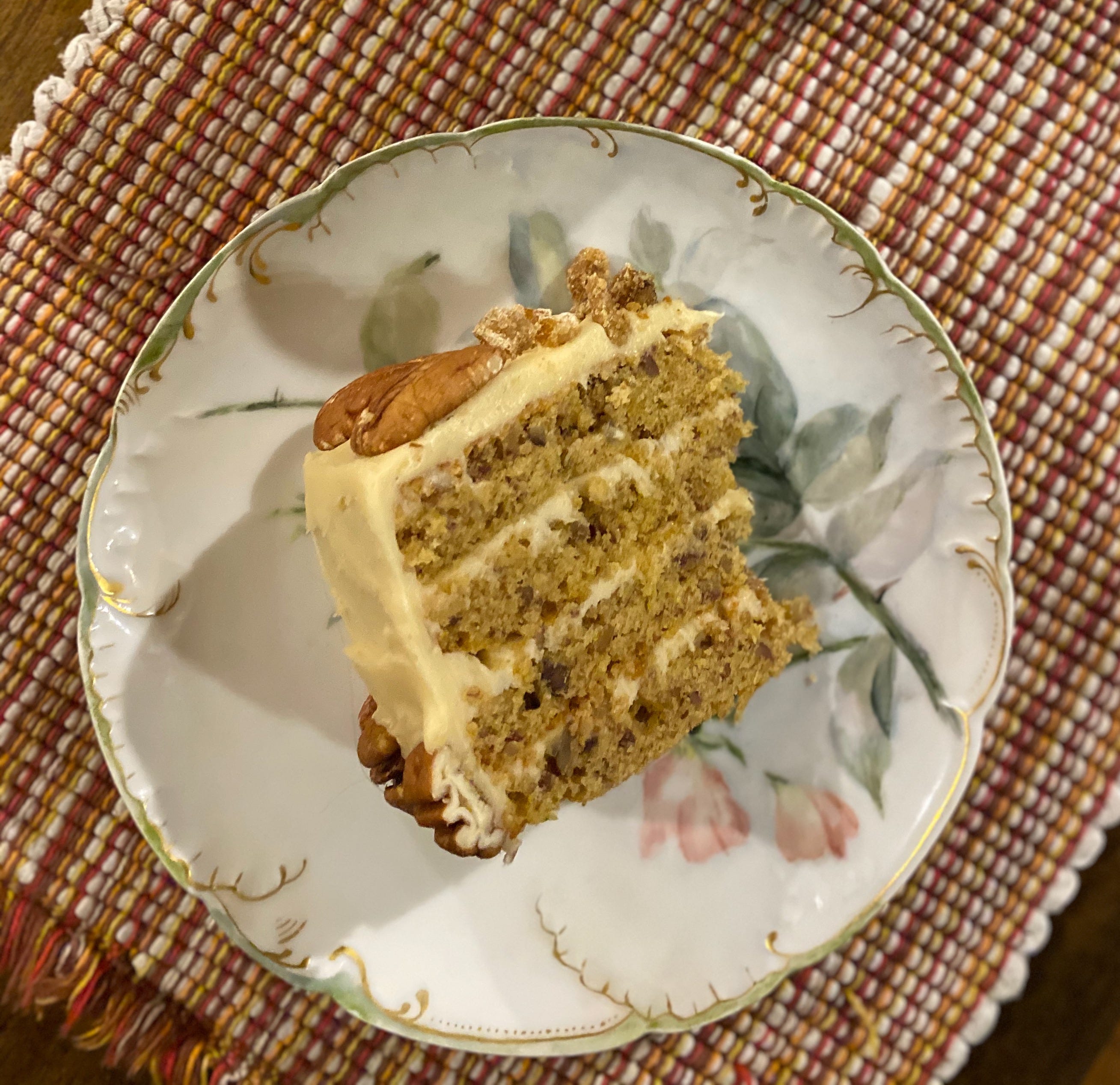 Slice of the above cake on a small plate painted with flowers. There are four layers, separated by frosting, with pecan halves on the top and side.