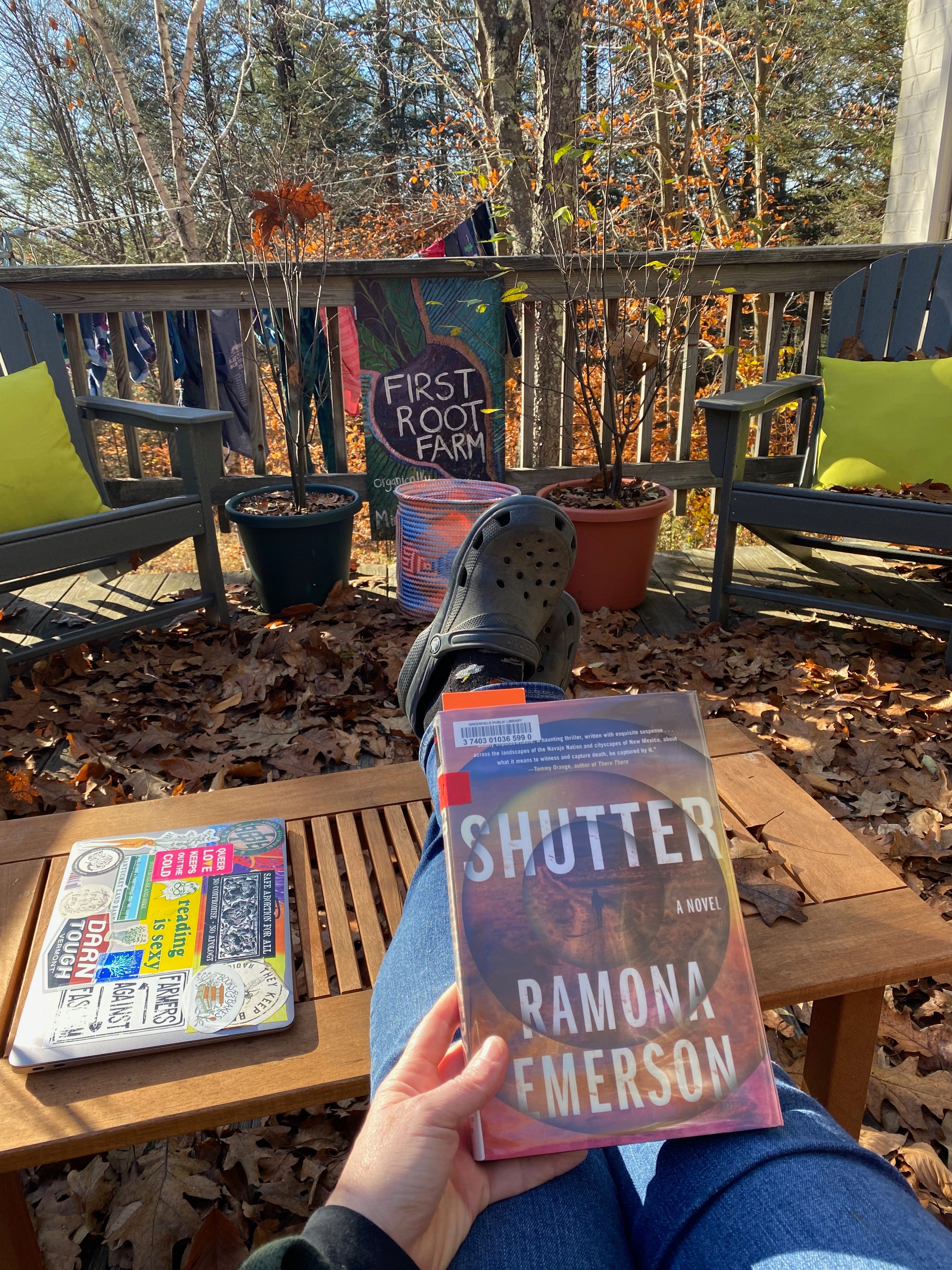 A view of my legs stretched out on a small wooden coffee table on my porch, which is covered in oak leaves and dappled with fall sunlight. I’m holding Shutter, and there’s a view of trees in the distance beyond the porch.
