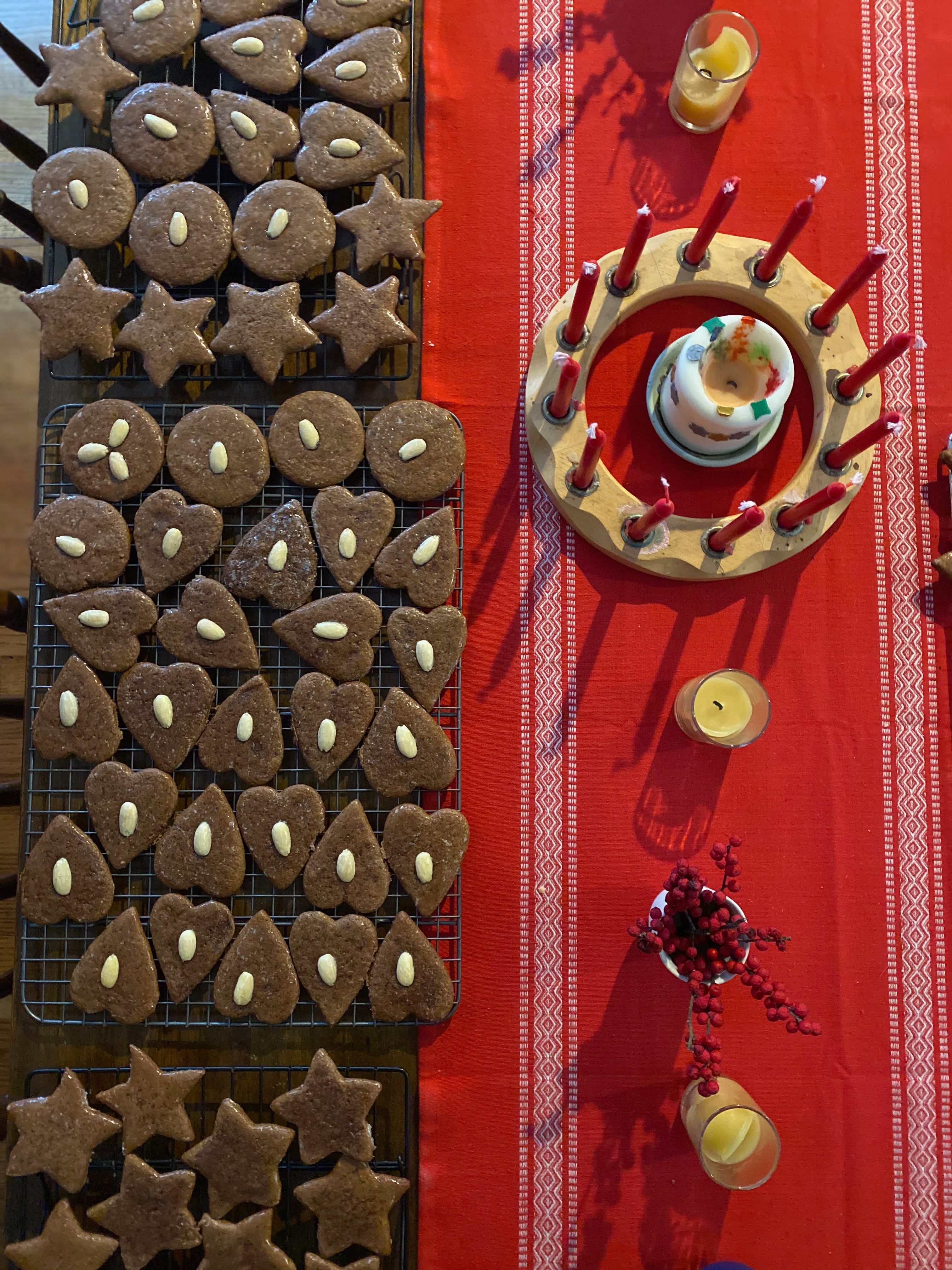 Three trays of round, heart, and star-shaped Lebkuchen, each with an almond in the center, sit on one side of a table. On the other side is a red tablecloth, with a round candleholder holing red advent candles, a small vase of winterberry, and three small beeswax candles in glass jars.