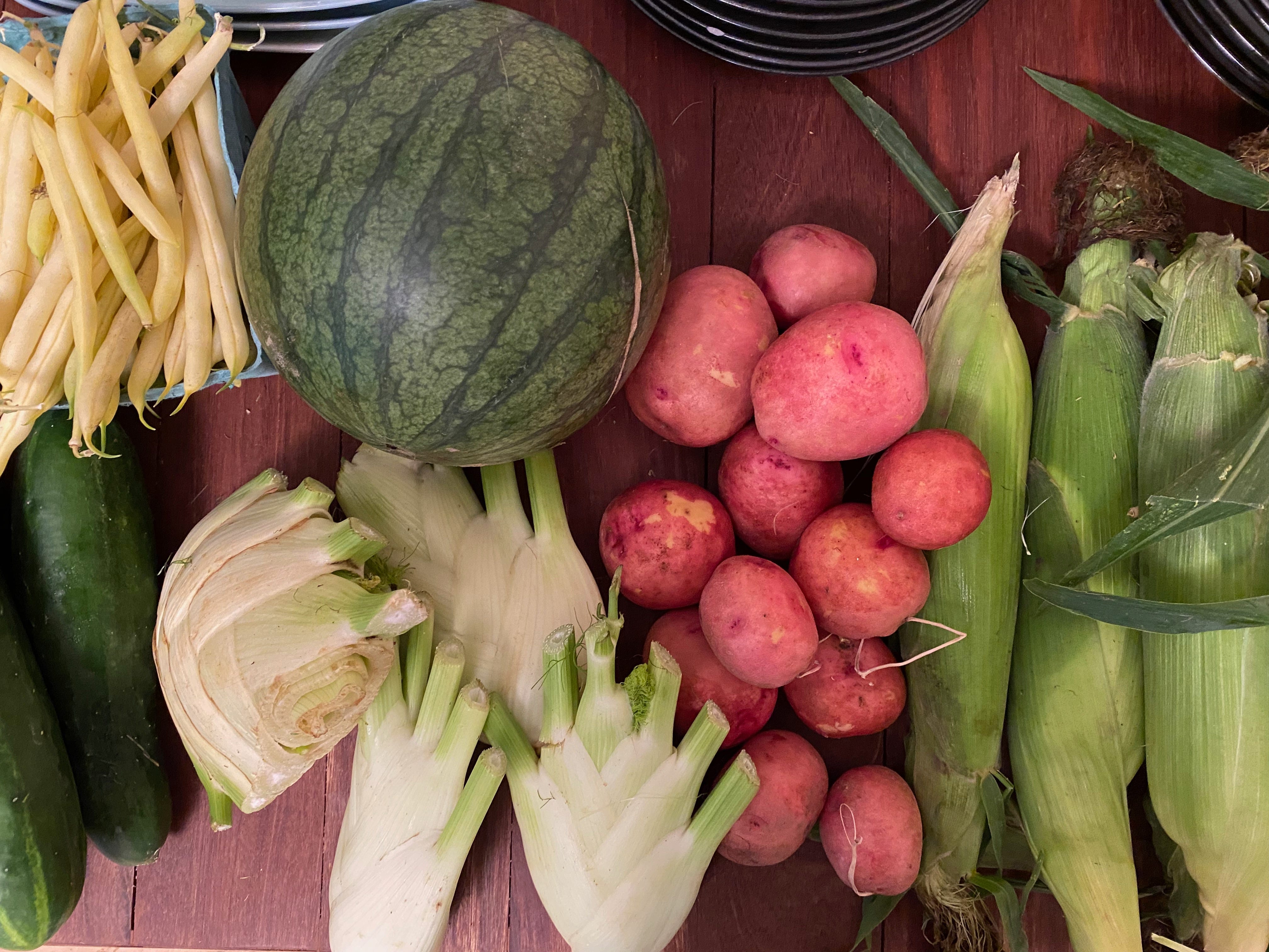 A pile of vegetables on a wooden counter: a quart of yellow beans, cucumbers, a watermelon, fennel, new read potatoes, and corn.