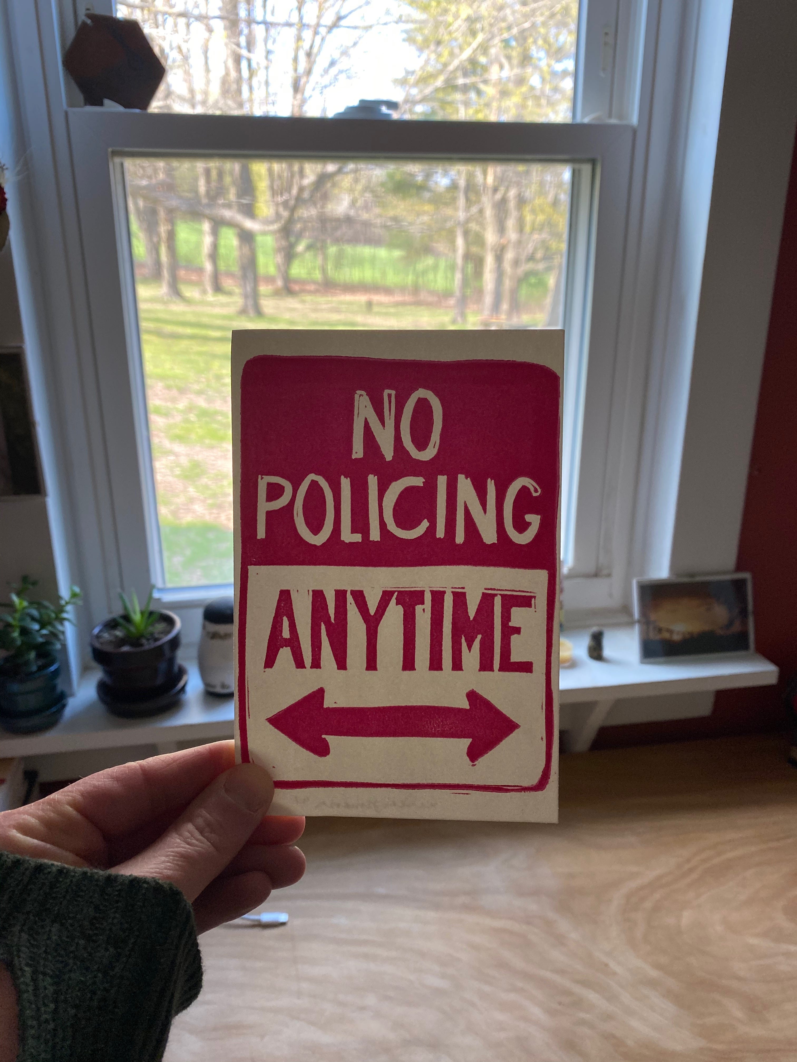 My hand holding a small linocut print featuring the words ‘No Policing Anytime’ and an arrow. It’s red and white, and made to look like a street sign. In the background is a view of trees and grass out the window.