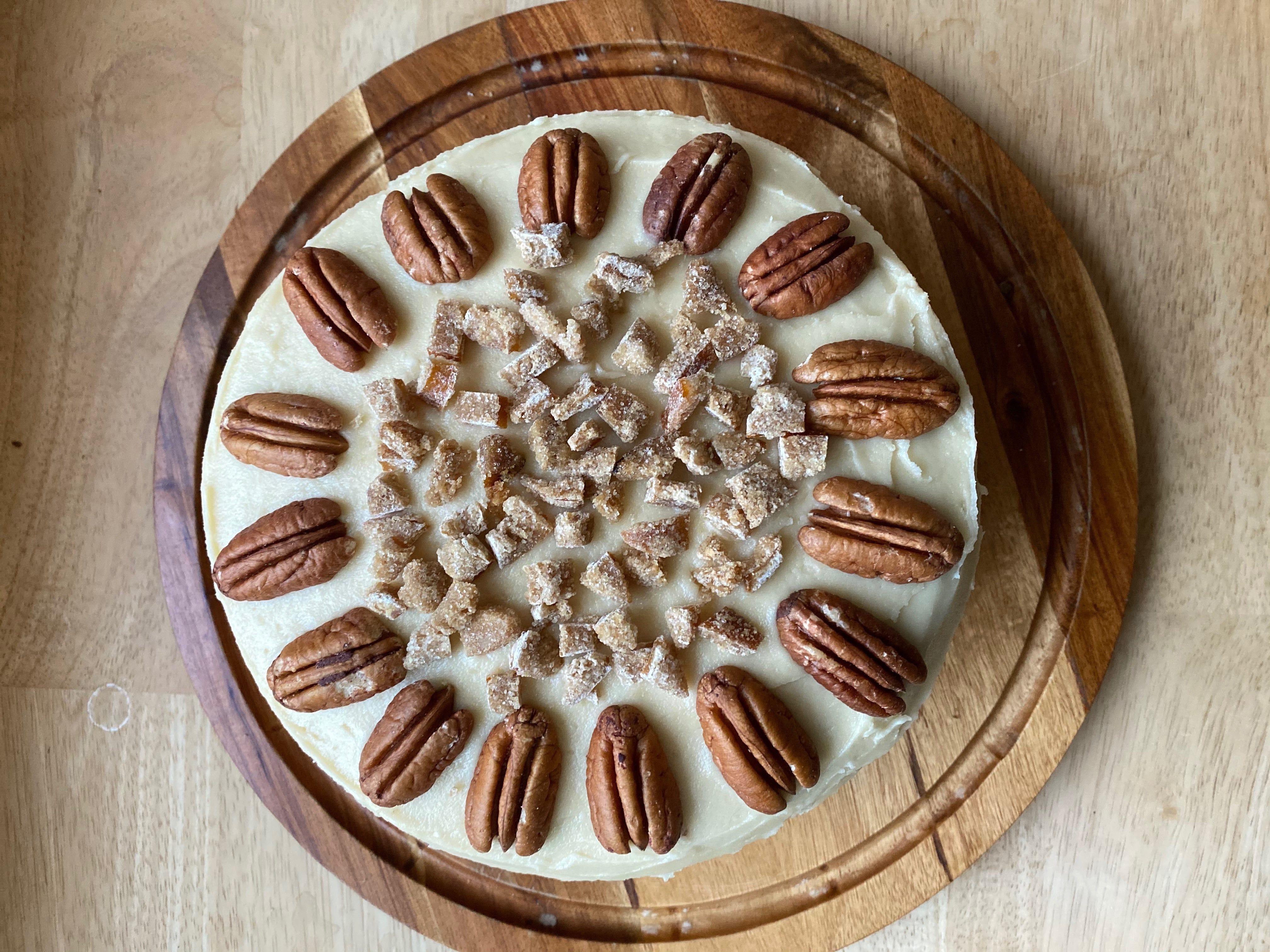 View from above of a round cake on a wooden board. It is covered in white frosting, with a ring of pecan halves arranged around the edges, and a pile of chopped candied orange peel in the center.