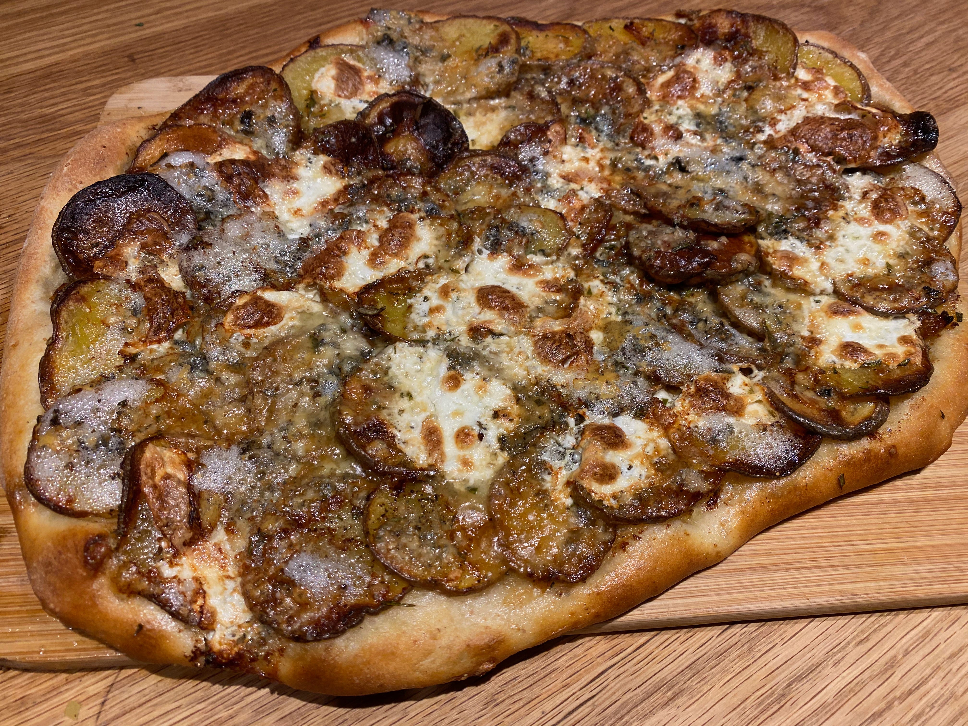 A potato pizza on a wooden cutting board. Thinly sliced potatoes are arranged in concentric circles, topped with browned, bubbly cheese.