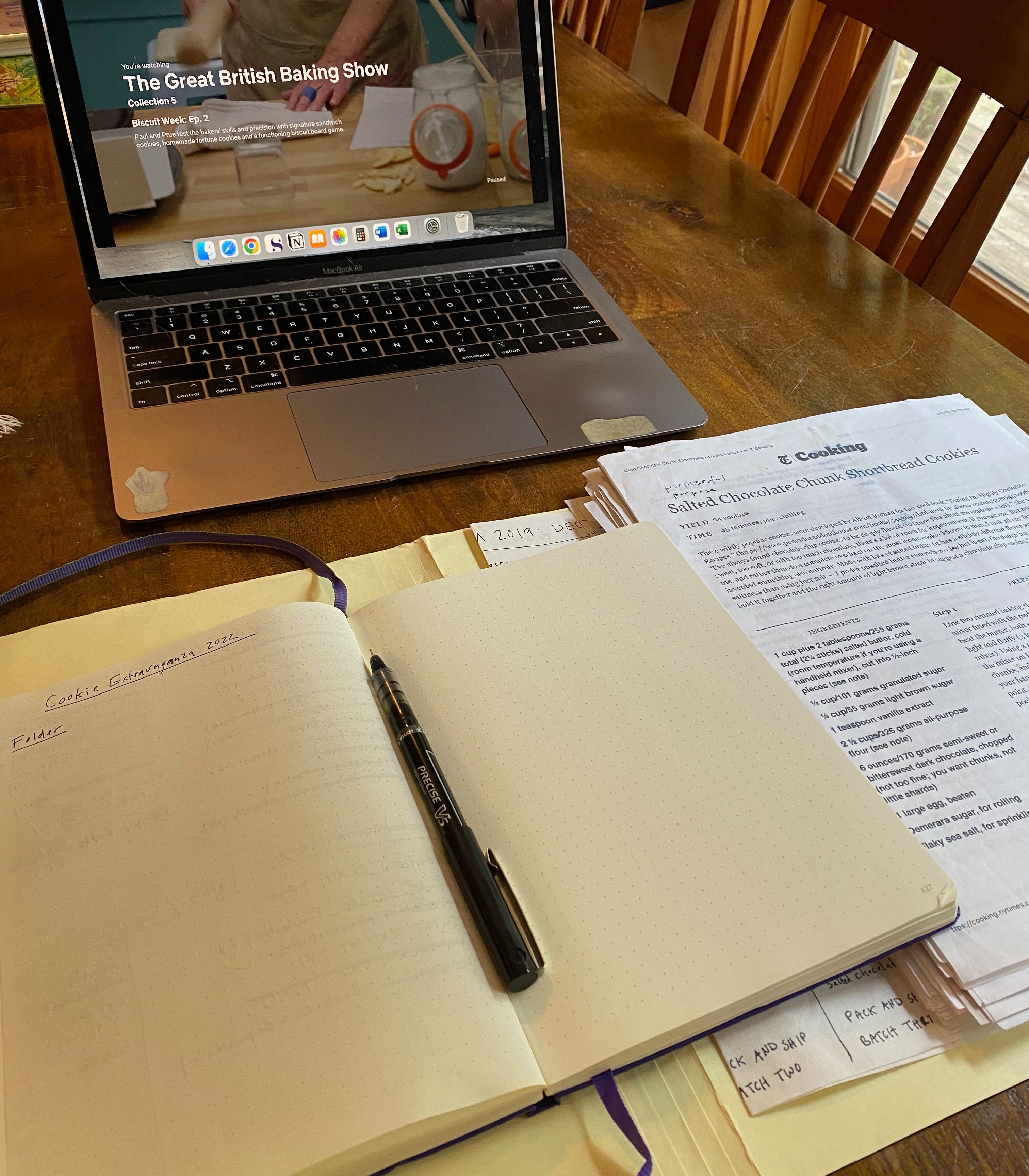 An open notebook on a wooden table next to a stack of printed-out cookie recipes. A laptop playing The Great British Bake Off sits behind the papers. 