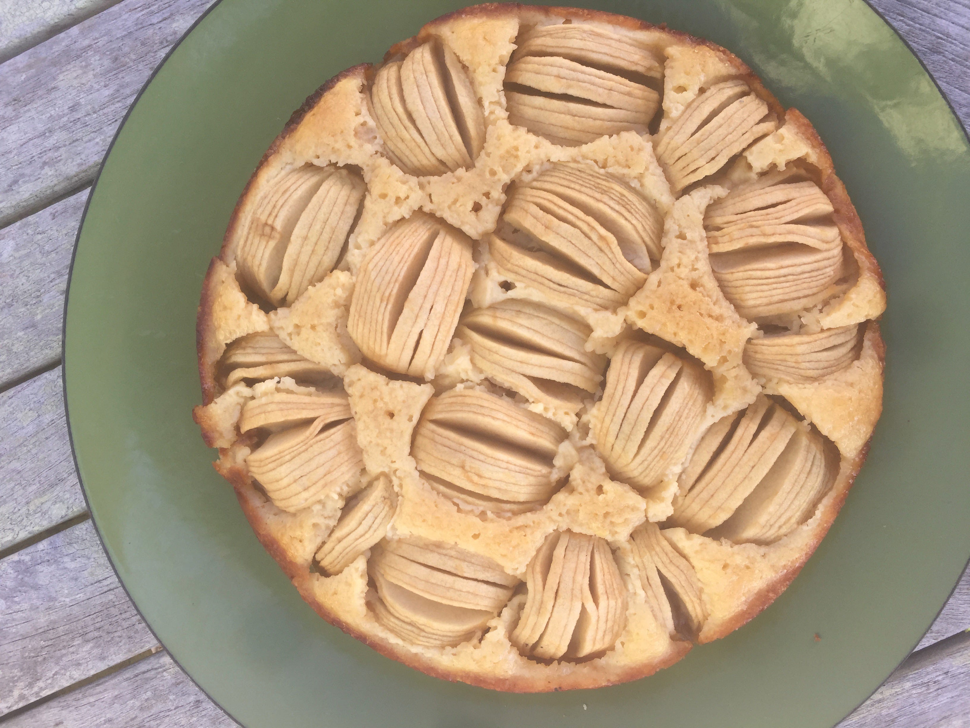 A round cake on a green platter. The cake is studded with apple halves, cut into thin wedges and fanned out into slices, sunken partway into the cake’s surface.