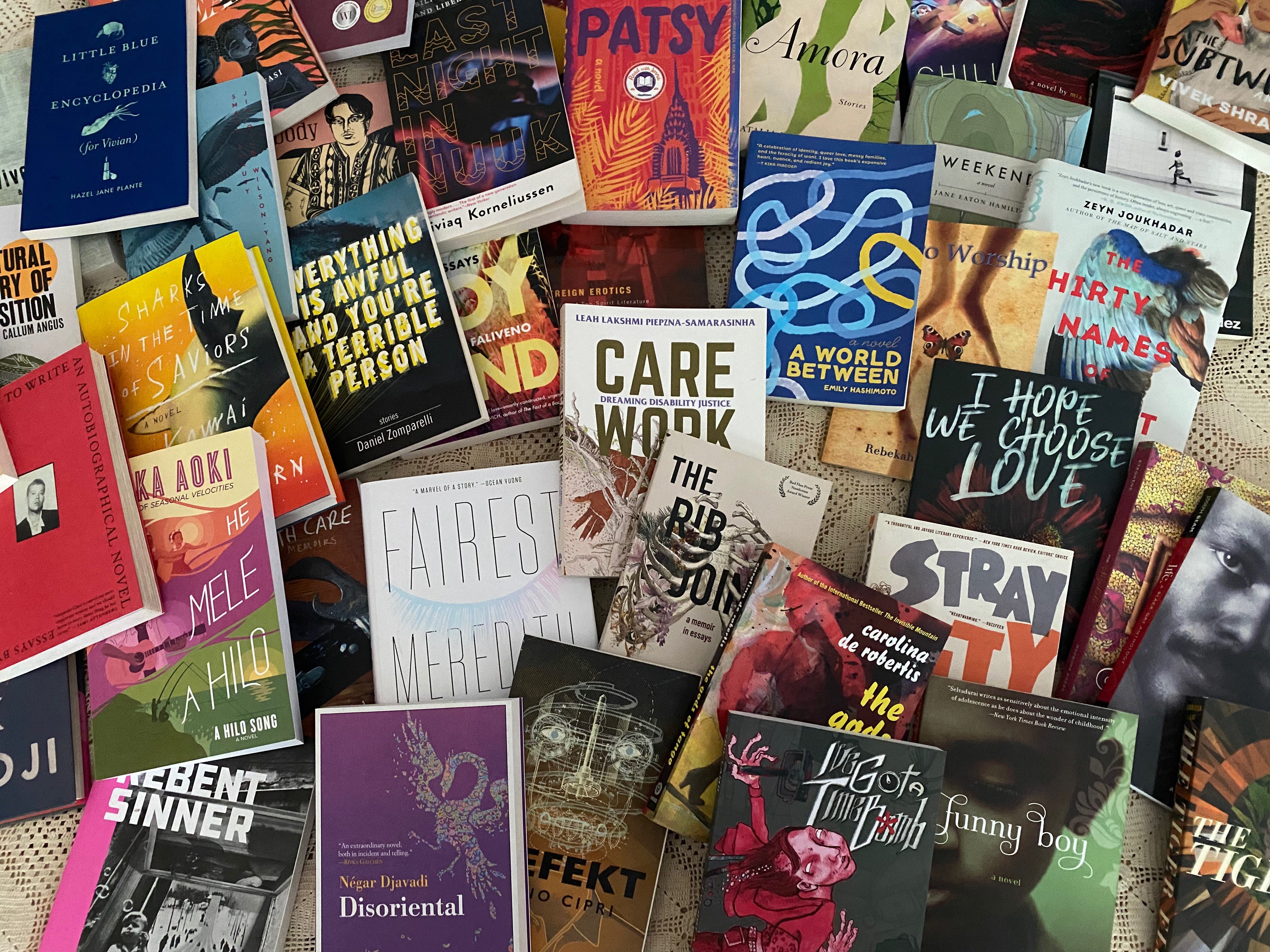 A messy pile of colorful, overlapping queer books on a lacy white bedspread: Little Blue Encyclopedia (for Vivian), Small Beauty, Last Night in Nuuk, Patsy, Amora, Chilling Effect, Weekend, The Subtweet, The Thirty Names of Night, Hero Worship, A World Between, Sovereign Erotics, Care Work, Everything is Awful and You're A Terrible Person, Sharks in the Time of Saviors, How to Write an Autobiographical Novel, He Mele A Hilo, Fairest, The Rib Joint, Defekt, Stray City, I Hope We Choose Love, The Gods of Tango, Funny Boy, Disoriental, I've Got A Time Bomb, Rebent Sinner, The Tiger Flu