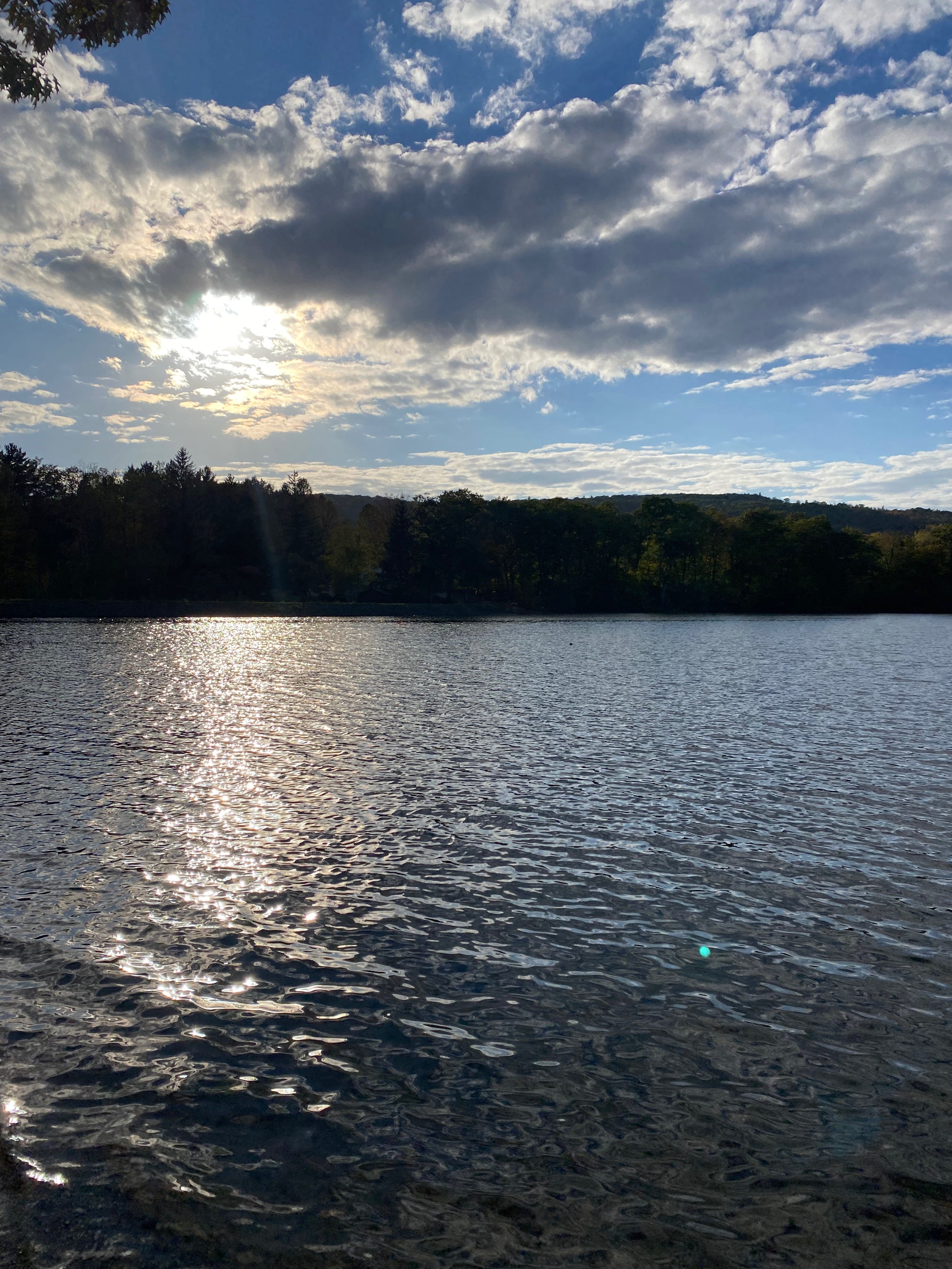 Ashfield Lake in the late afternoon. The sun, hidden behind a bank of clouds, casts a dazzling band of light across the dark, rippled water.