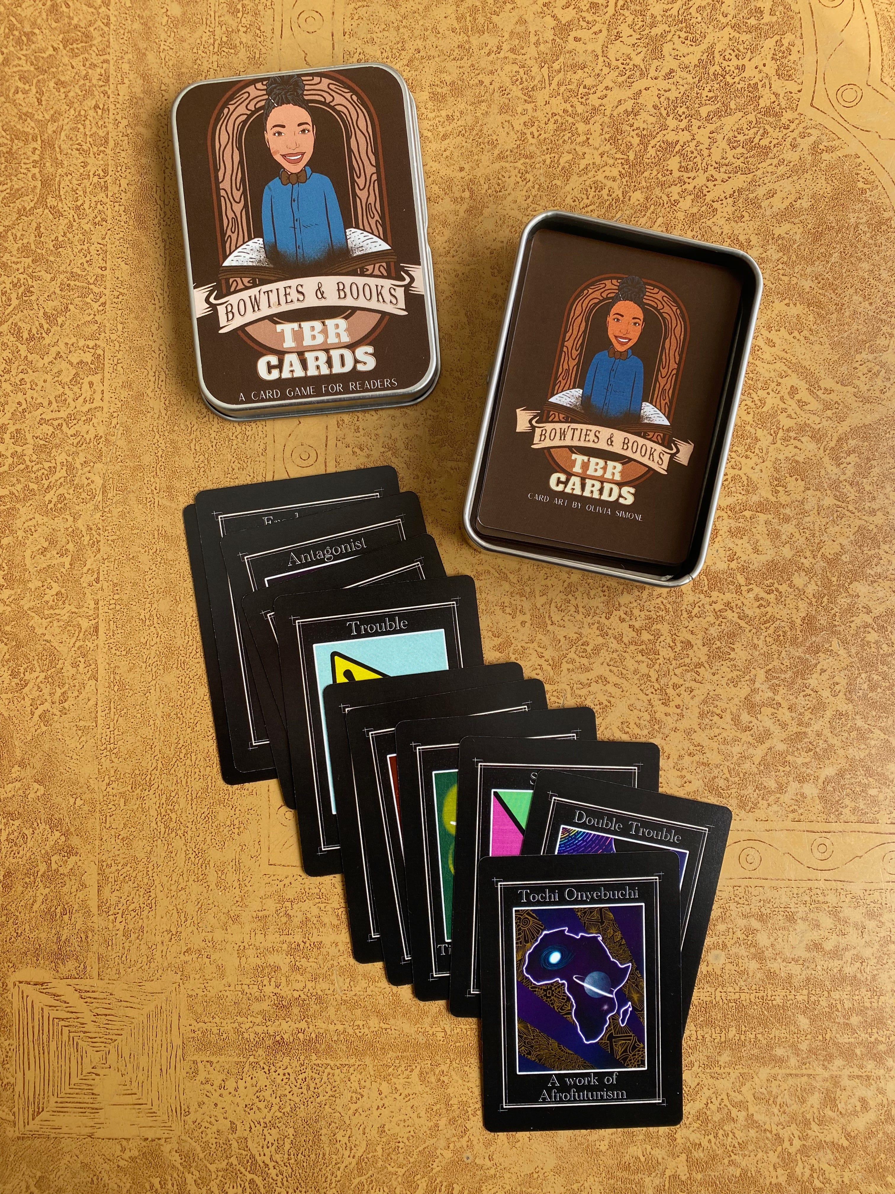 A small open tin of playing cards. The cards in the tin, as well as the cover of the tin, show a picture of a Black person and the words “Bowties & Books TBR Cards: A Card Game for Readers”. Several cards, showing colorful illustrations and prompts, are fanned out below the tin.