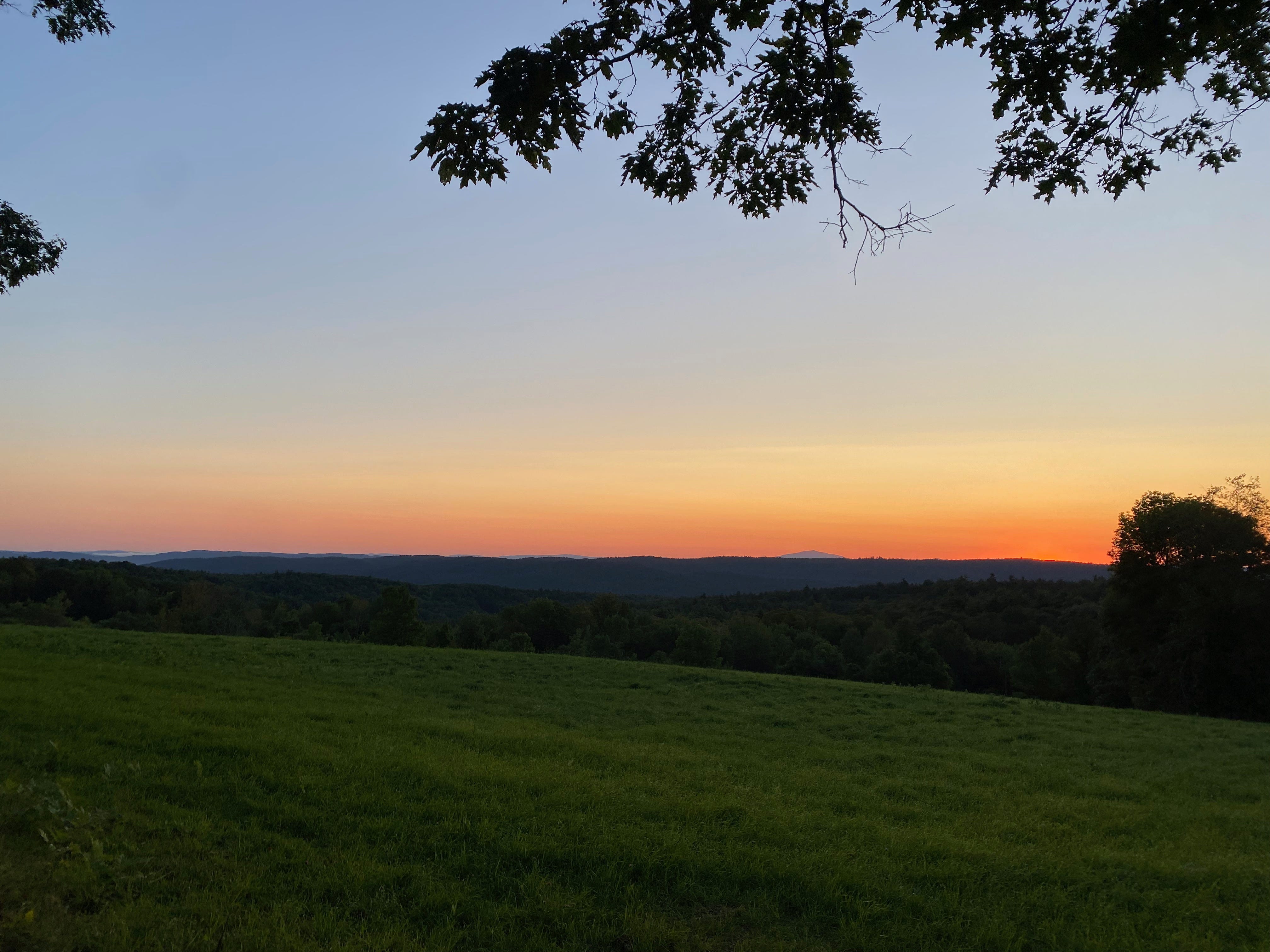Sunrise over a green hilltop pasture. Behind the distant hills and mountains, the horizon is deep orange and pink.