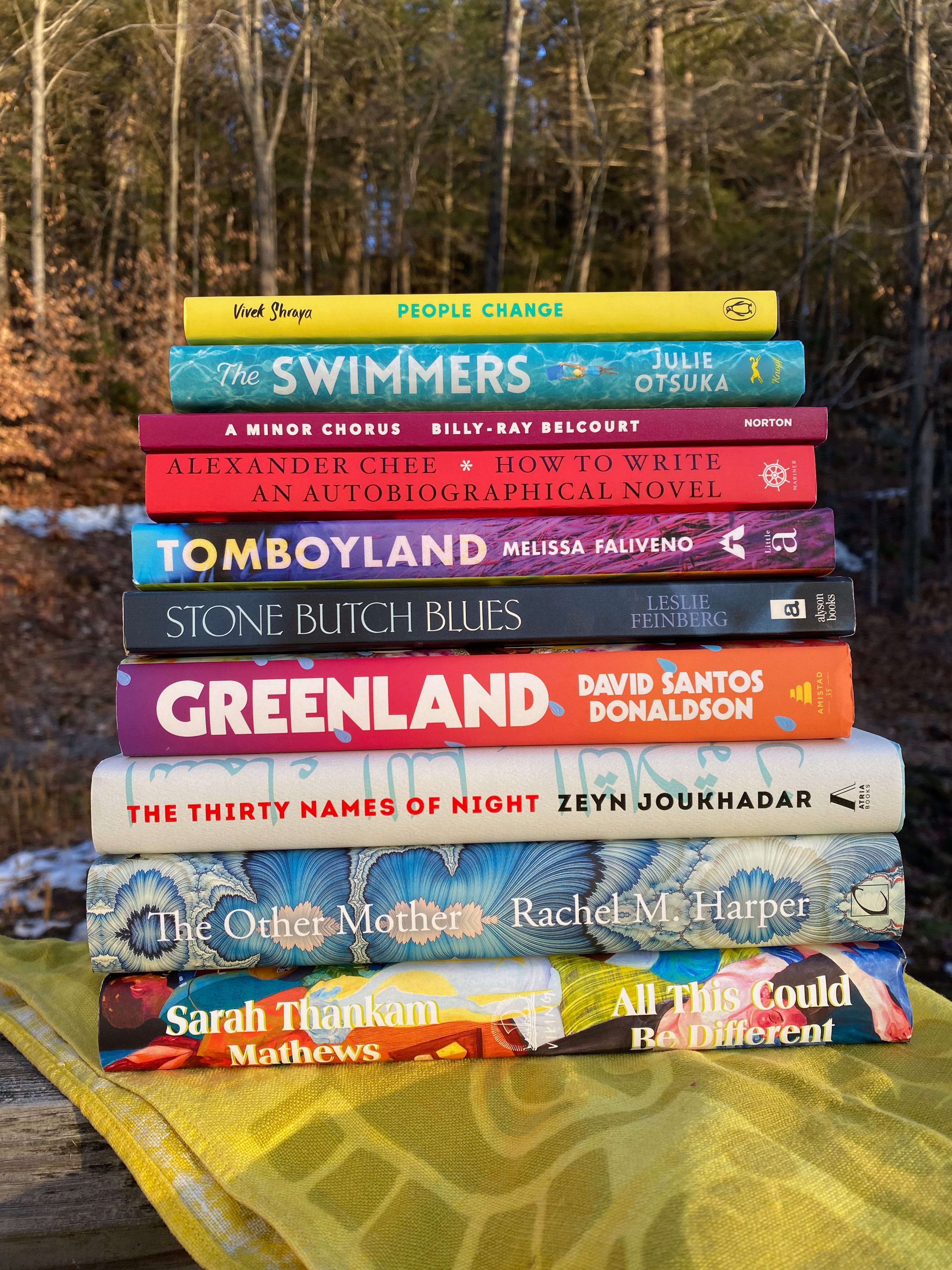 A stack of books on a yellow cloth on porch railing in the sun, with trees in the background. The books: All This Could Be Different, The Other Mother, The Swimmers, A Minor Chorus, Greenland, The Thirty Names of Night, Tomboyland, How to Write An Autobiographical Novel, Stone Butch Blues, and People Change.