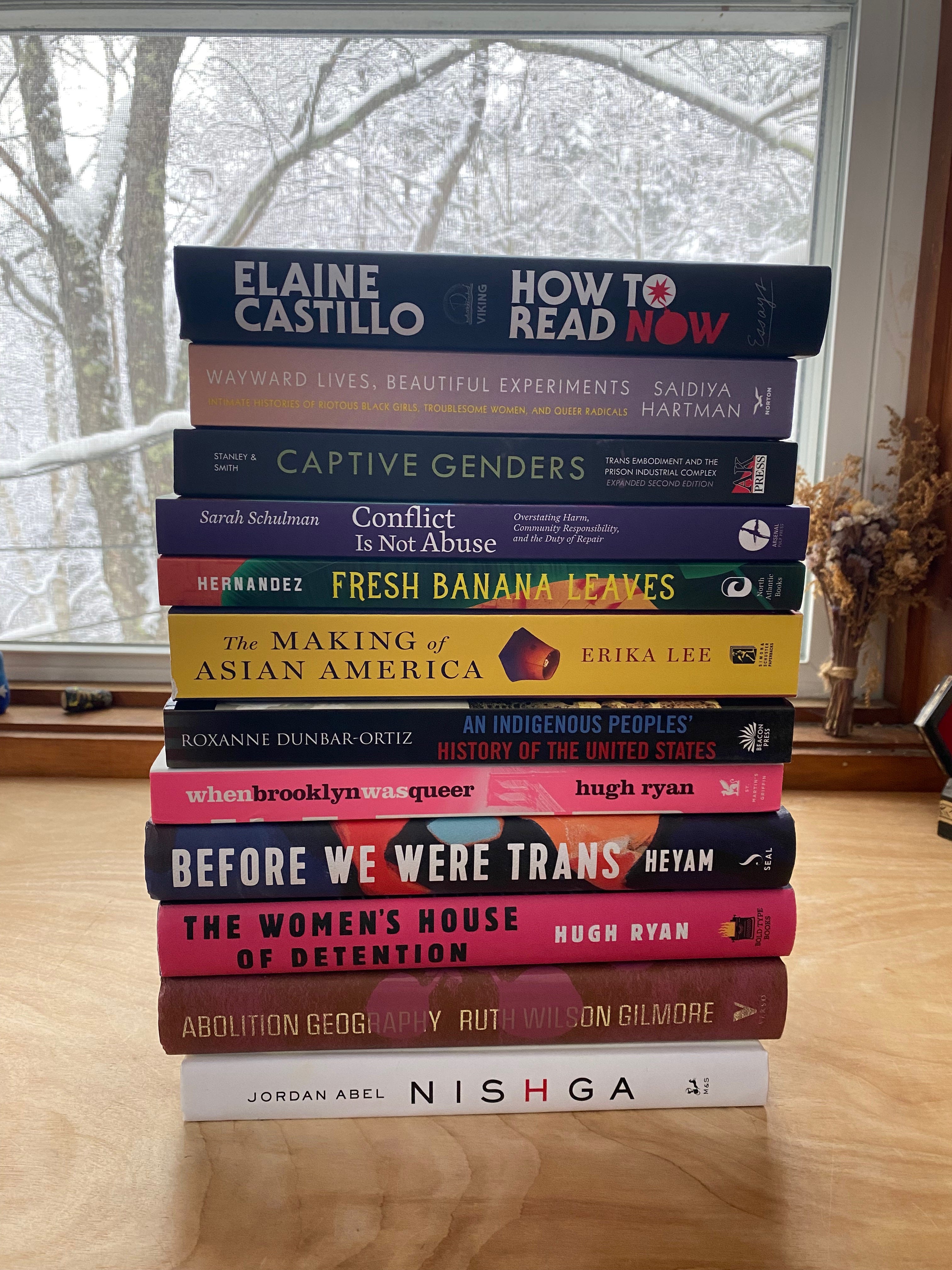 A big stack of books on a desk in front of a window. The books are: Abolition Geography , The Women's House of Detention, Before We Were Trans,  When Brooklyn Was Queer,An Indigenous Peoples' History of the United States, The Making of Asian America, Fresh Banana Leaves, Conflict is Not Abuse, Captive Genders, Wayward Lives, Beautiful Experiments, How to Read Now and Nishga.