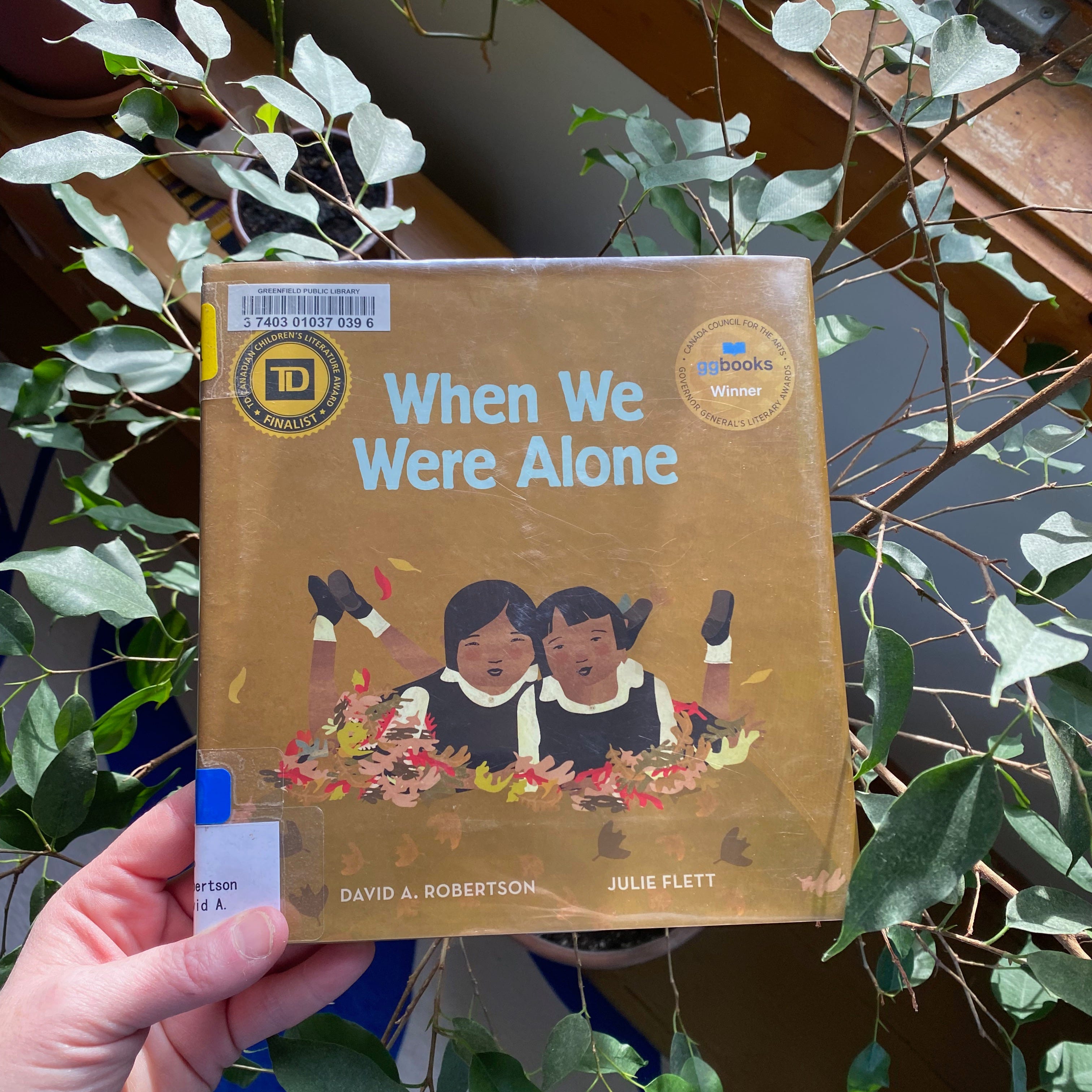 I’m holding up When We Were Alone in front of a ficus tree under a window.