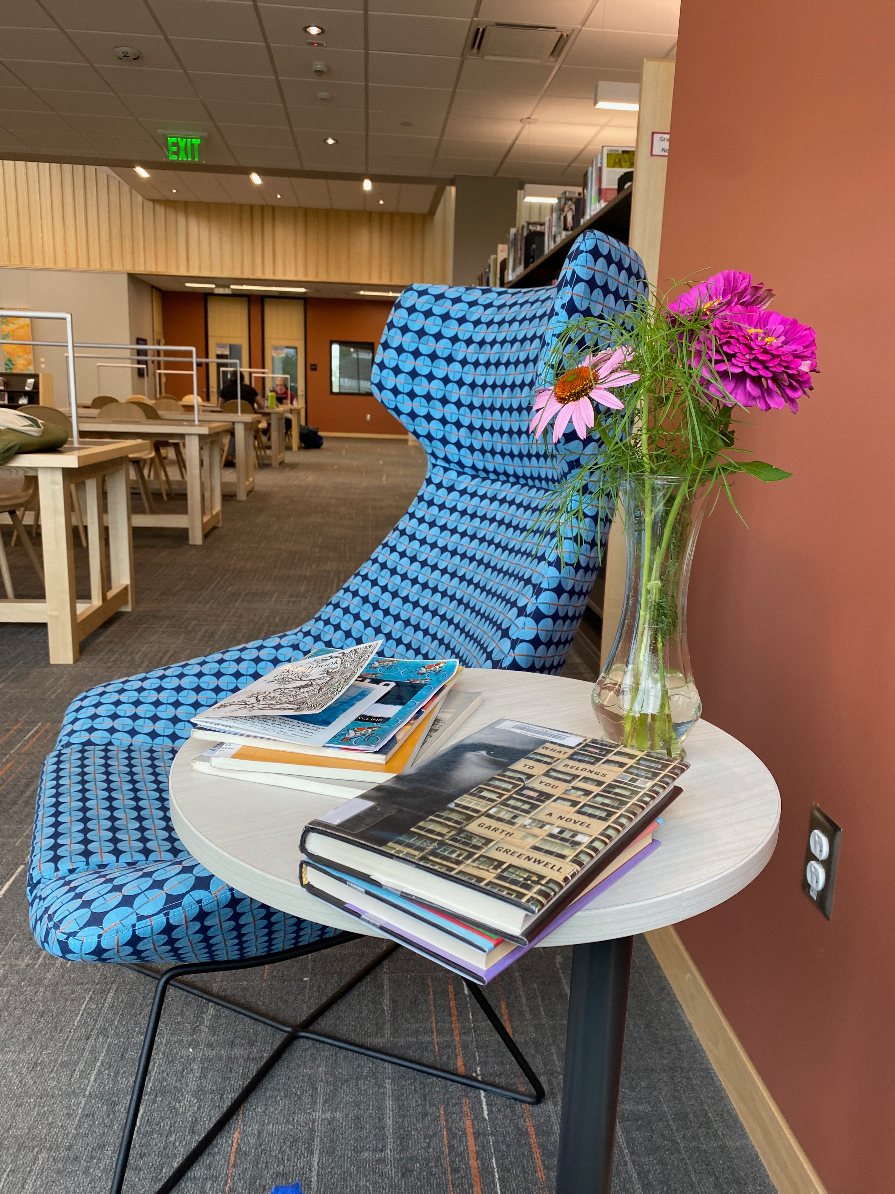 A stack of books and notebooks on a small table next to a blue armchair; rows of library tables are visible in the background.